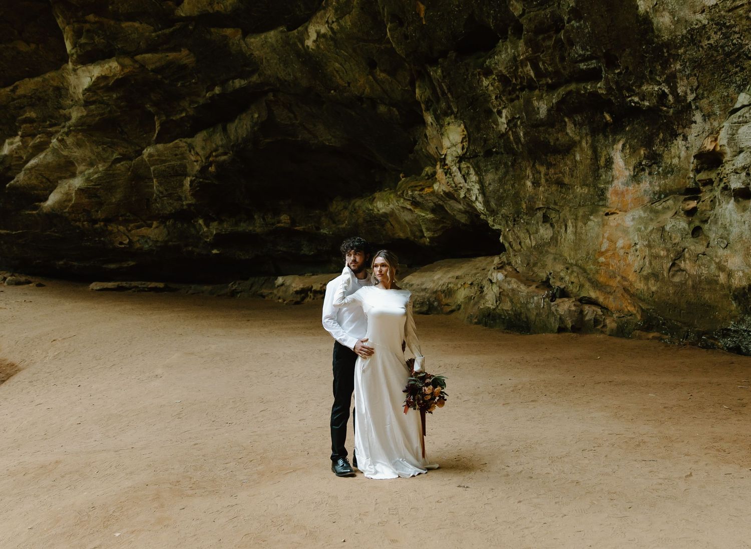 A couple shares an intimate moment during their wedding photoshoot inside a dramatic cave setting with sandy floor.
