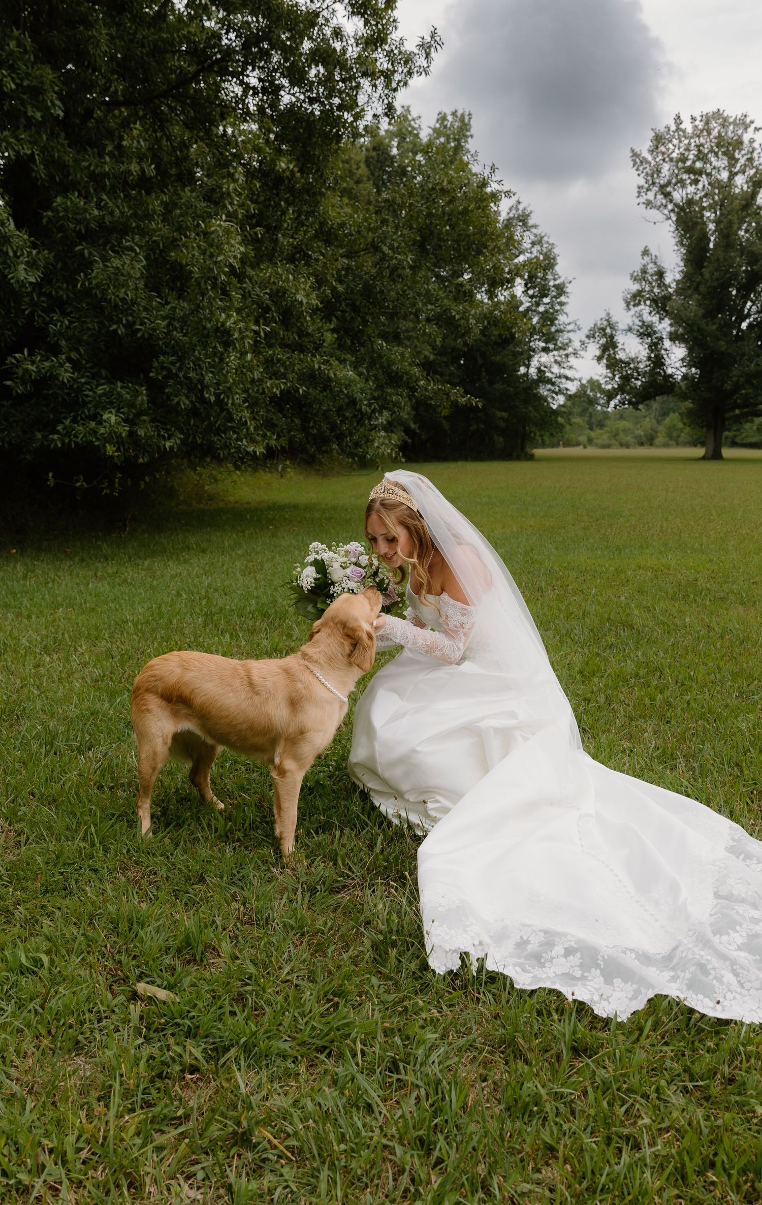 Bride in white gown and veil kneels on green grass, sharing her bouquet with a golden dog outdoors.