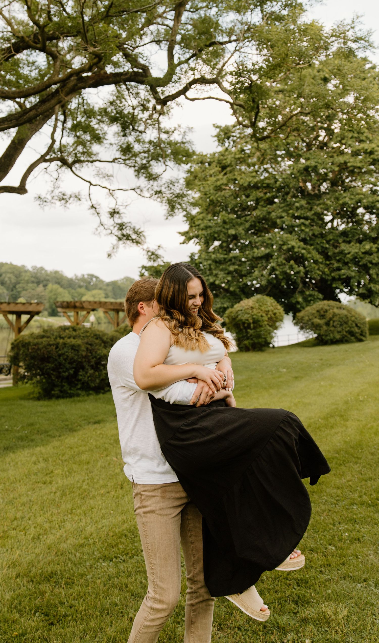 A romantic couple embraces on a grassy lawn beside trees and water on a sunny afternoon.