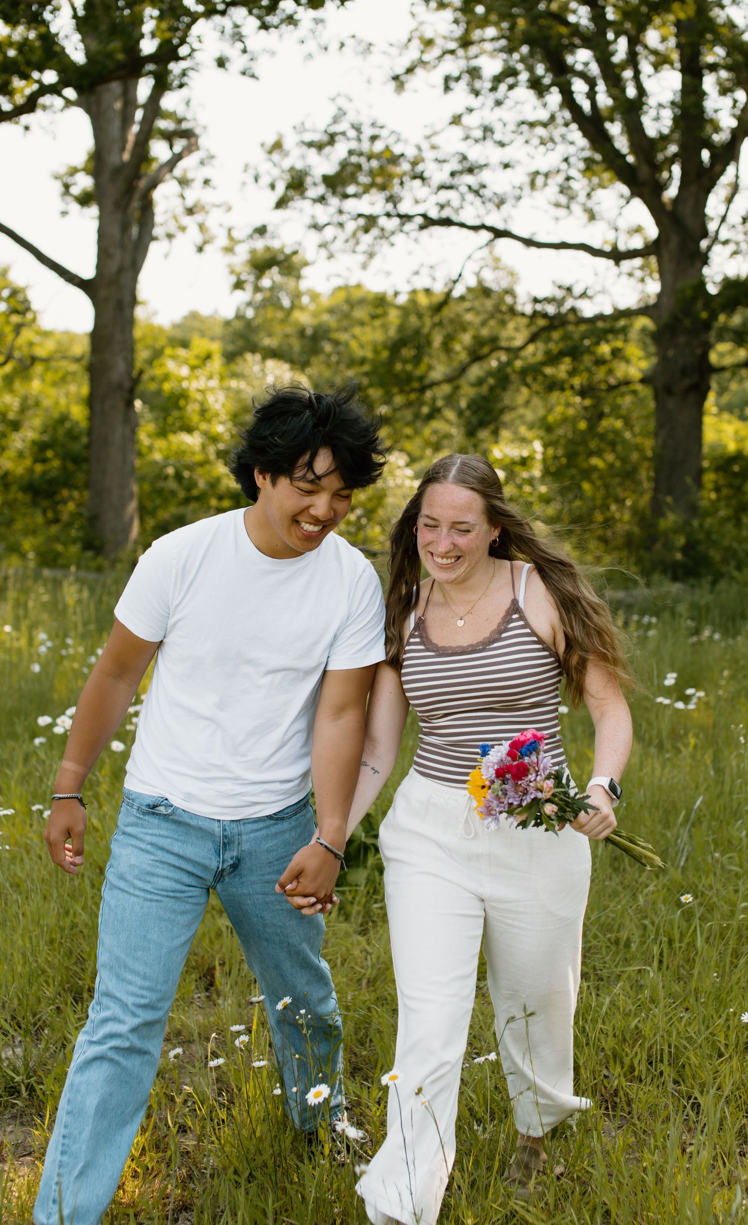 Joyful couple walking through wildflower meadow at sunset, holding hands while laughing together in nature.