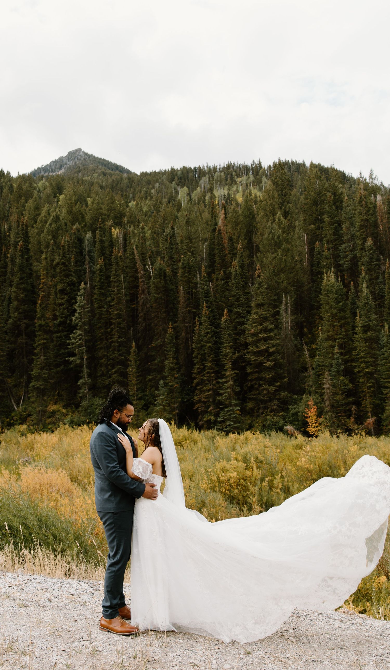 A wedding couple embraces against a stunning mountain backdrop with evergreen forests and flowing white dress veil.