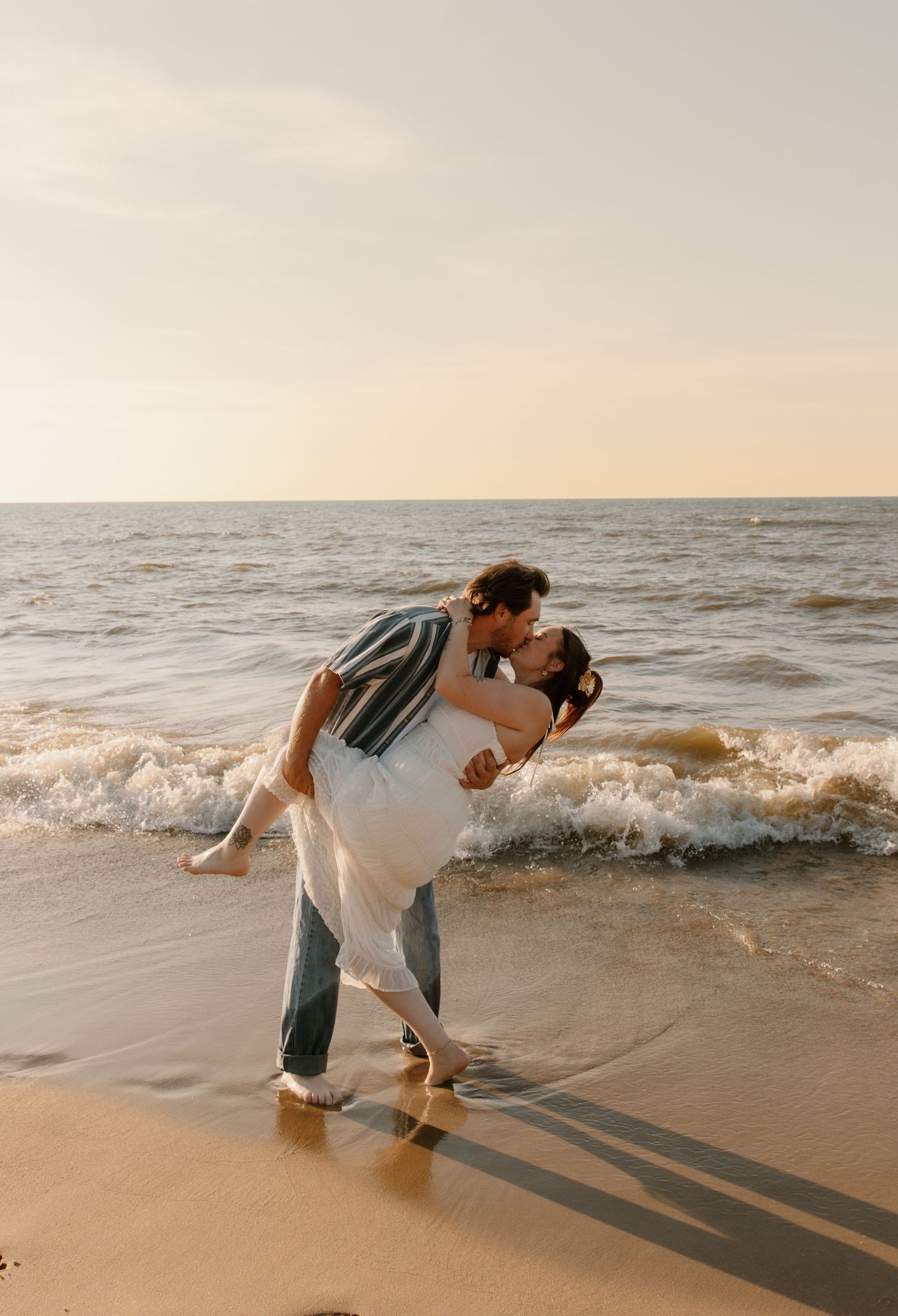 Couple sharing a romantic kiss on a sandy beach at sunset, with ocean waves crashing behind them.