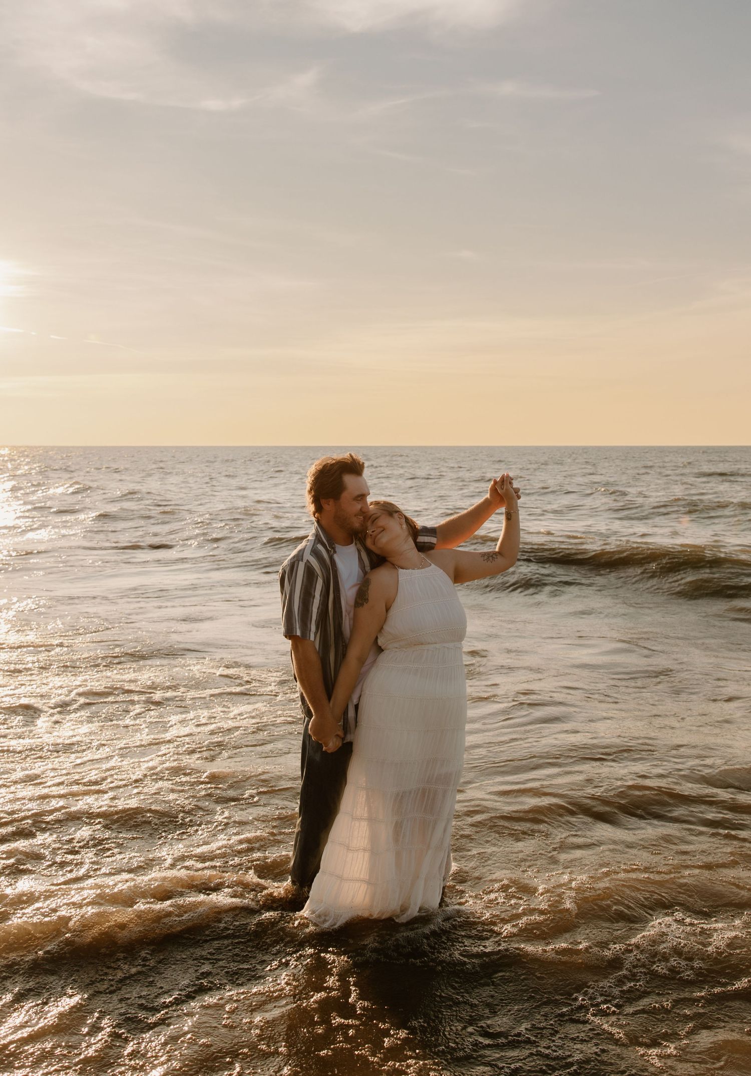 Couple dancing in ocean waves at sunset, woman in white dress, romantic beach wedding photo.