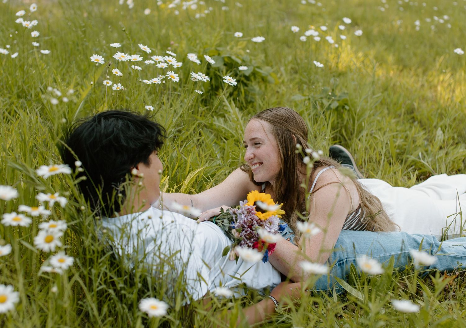Two people lying in a summer meadow filled with white daisies and holding wildflowers while enjoying a peaceful moment.