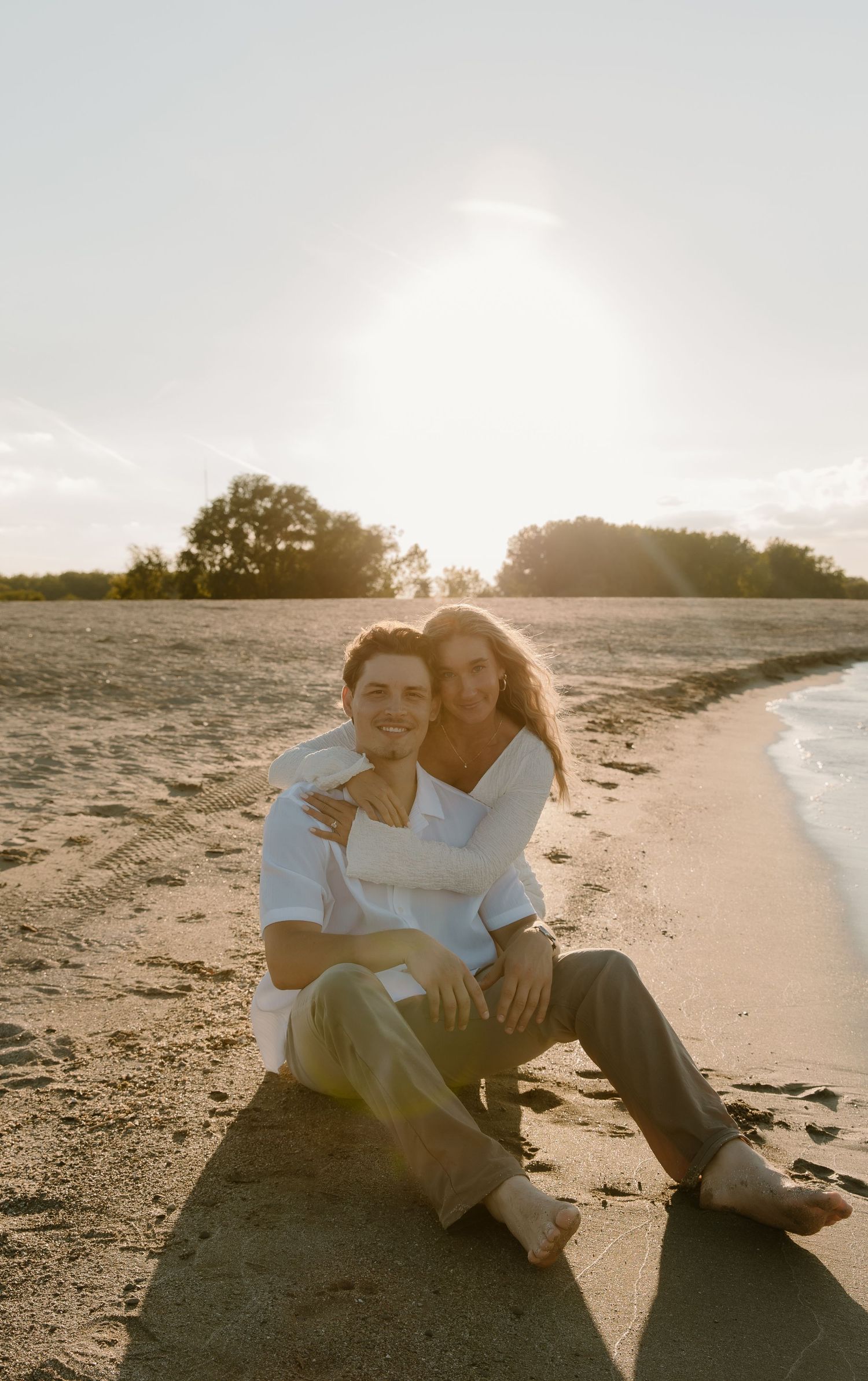 A couple sits together on a sandy beach at golden sunset, smiling warmly with trees and ocean in the background.