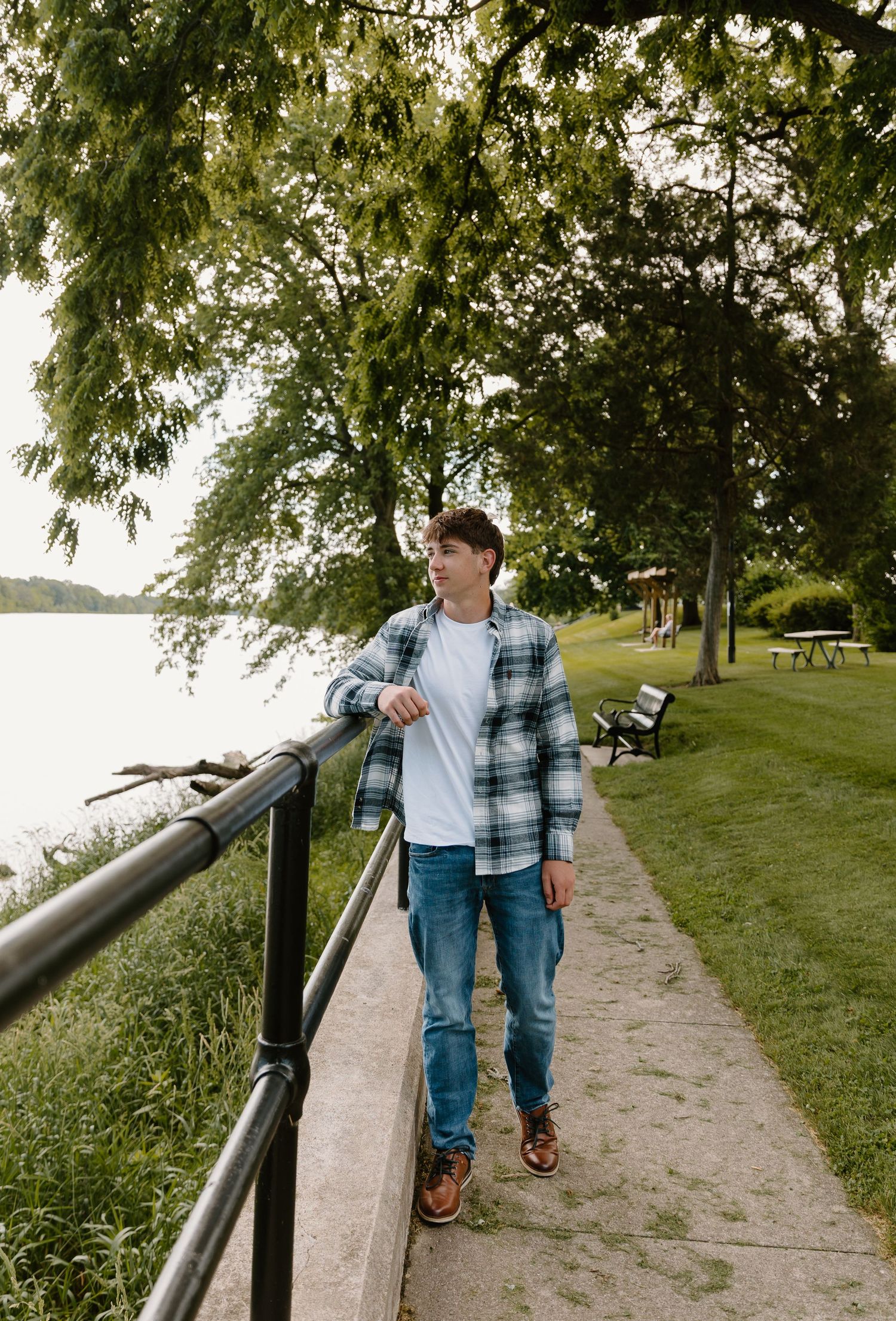 Young man in plaid shirt and jeans leaning on railing by a lake in a lush green park on a cloudy day.