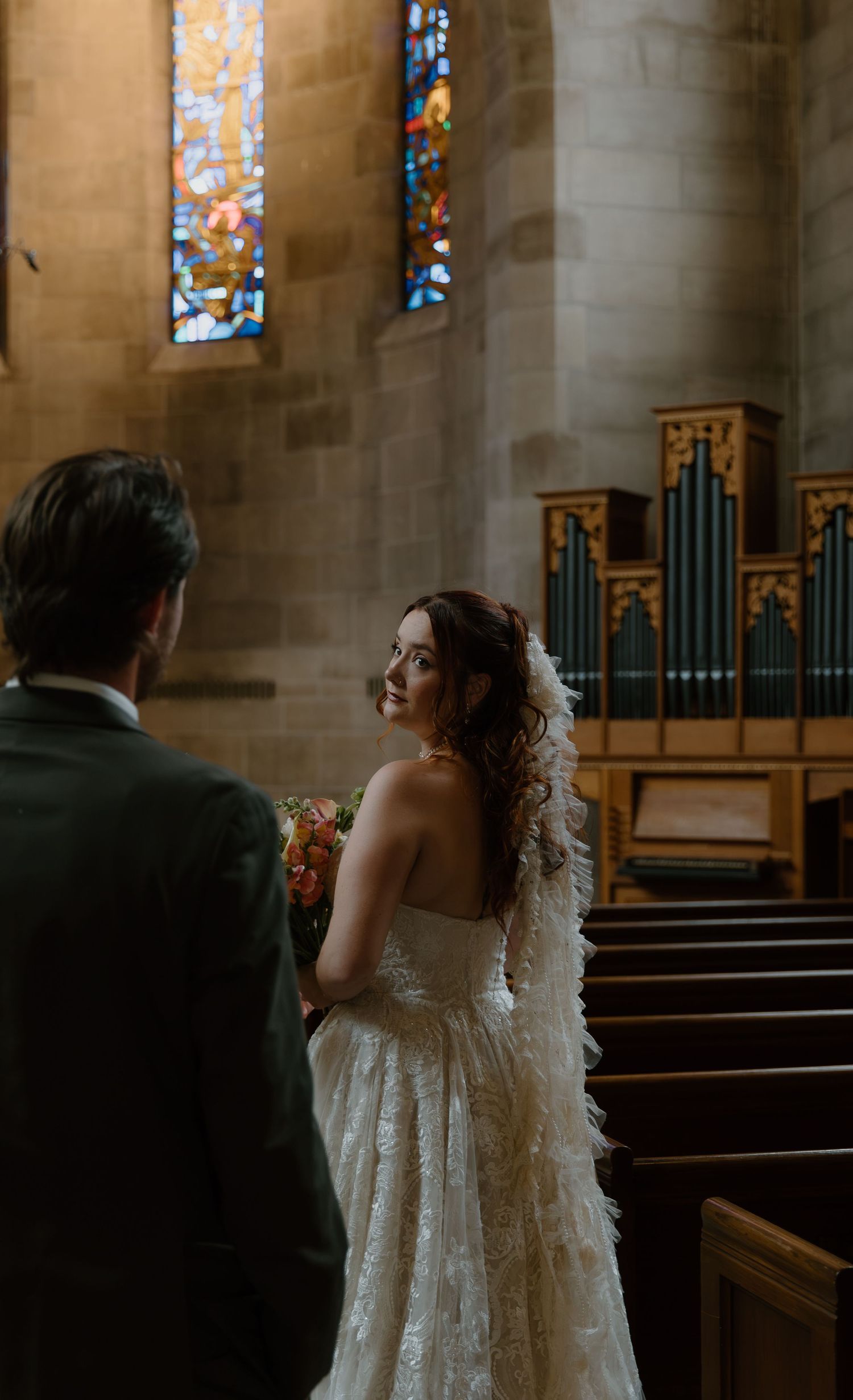Bride in lace gown smiles at groom inside a church with stained glass windows and pipe organ.