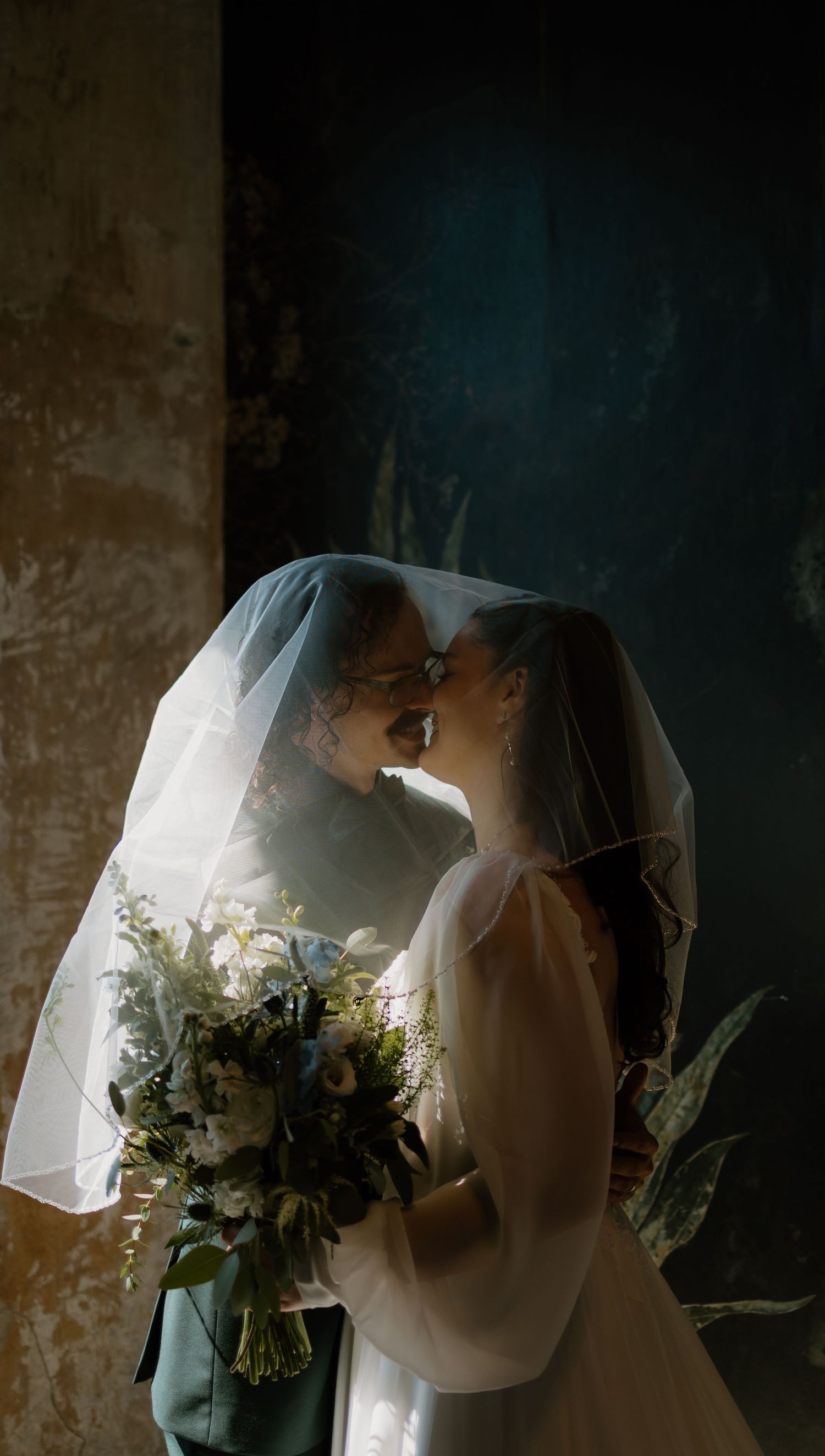 A bride and groom share an intimate moment under a sheer veil, with the bride holding a lush white and green bouquet.