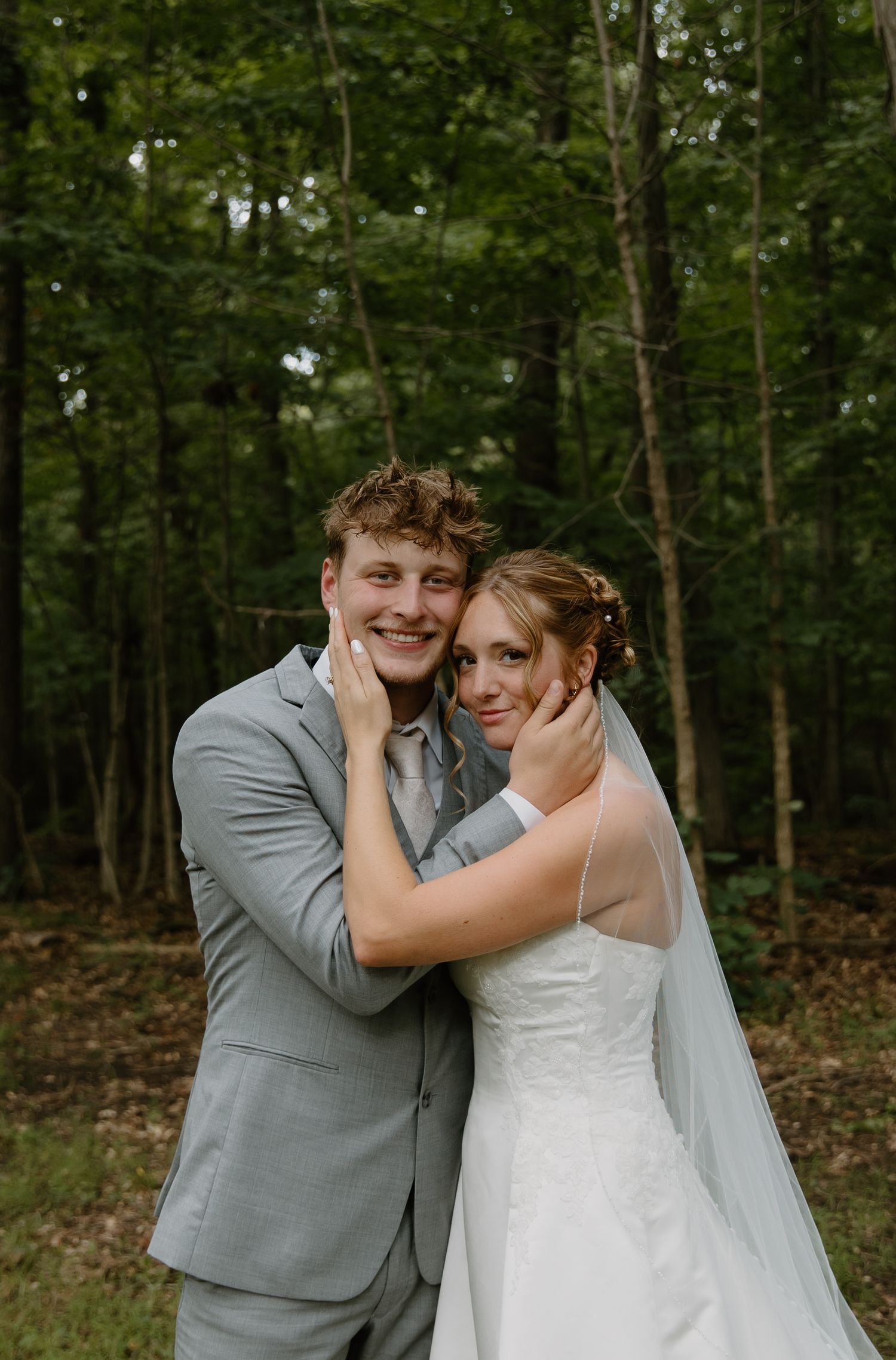 A smiling groom in a gray suit embraces his bride in a white gown and veil outdoors in a lush green forest setting.