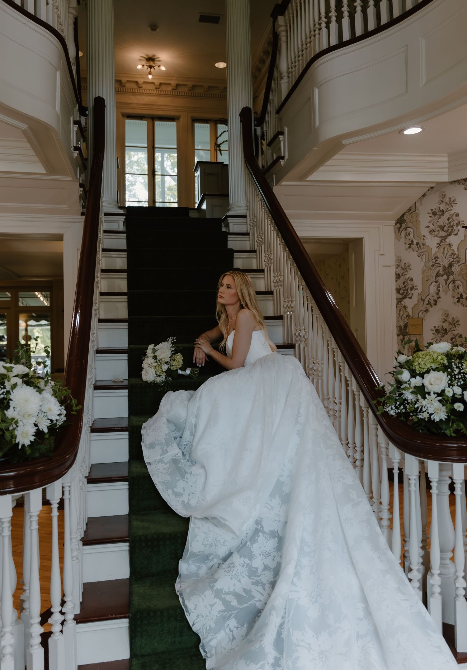 Bride in dramatic lace ballgown poses on grand curved staircase adorned with white floral arrangements.