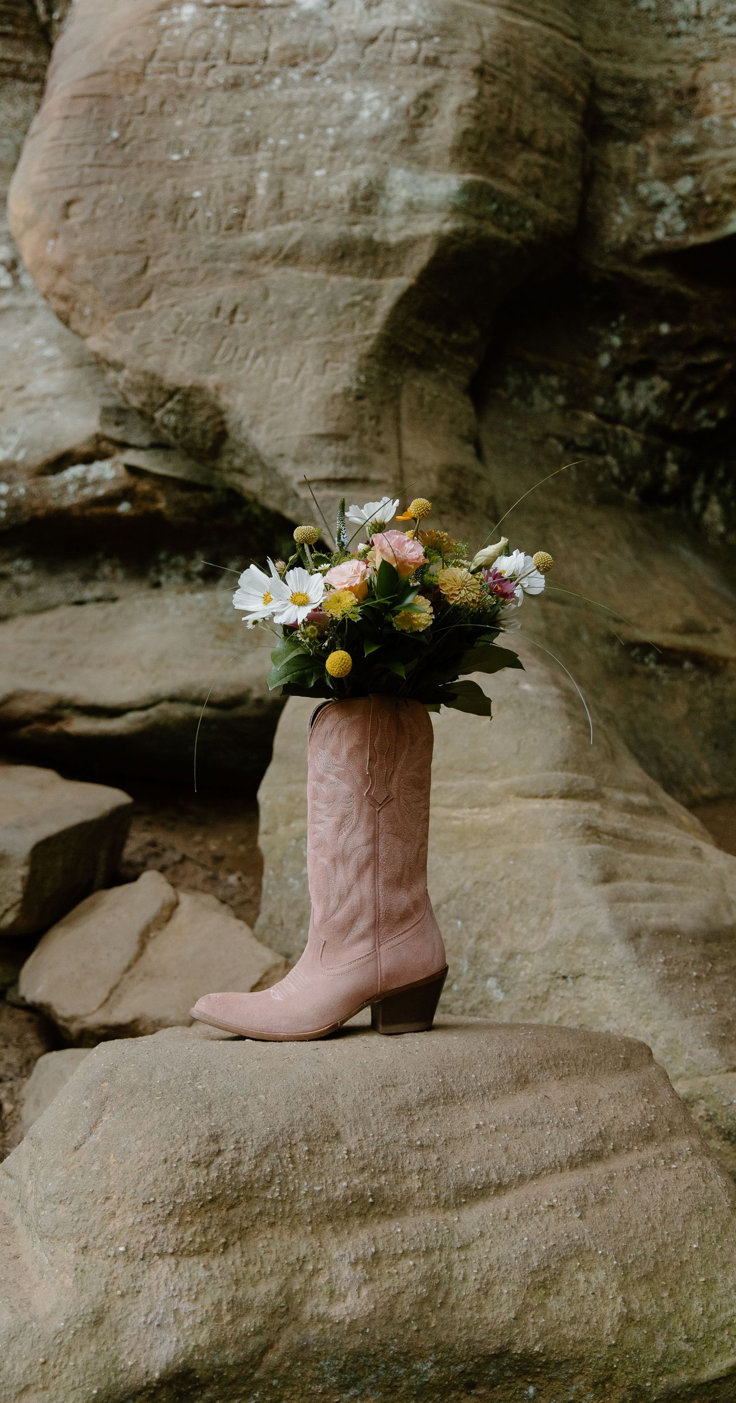 Pink cowboy boot filled with fresh wildflowers sits on a weathered rock surface in natural light.