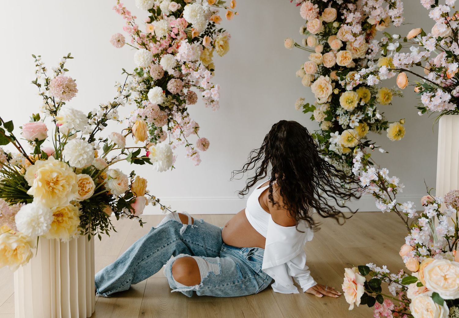Woman in white crop top and ripped jeans sitting on floor surrounded by lush pink and white floral arrangements.