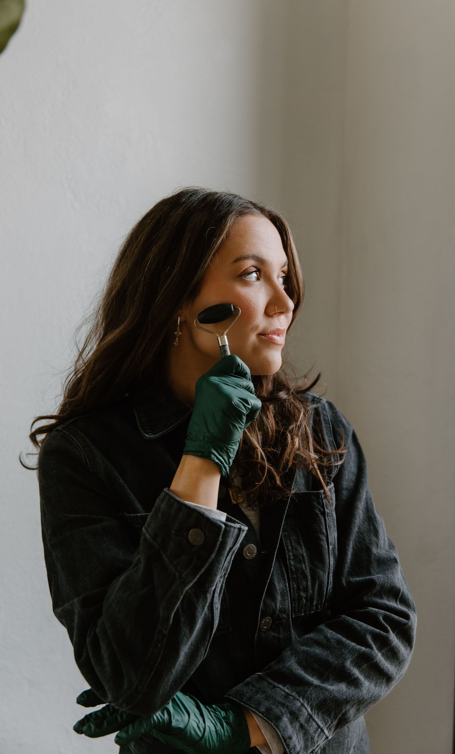 Woman in black denim jacket using a jade roller facial tool while wearing green gloves near a window.