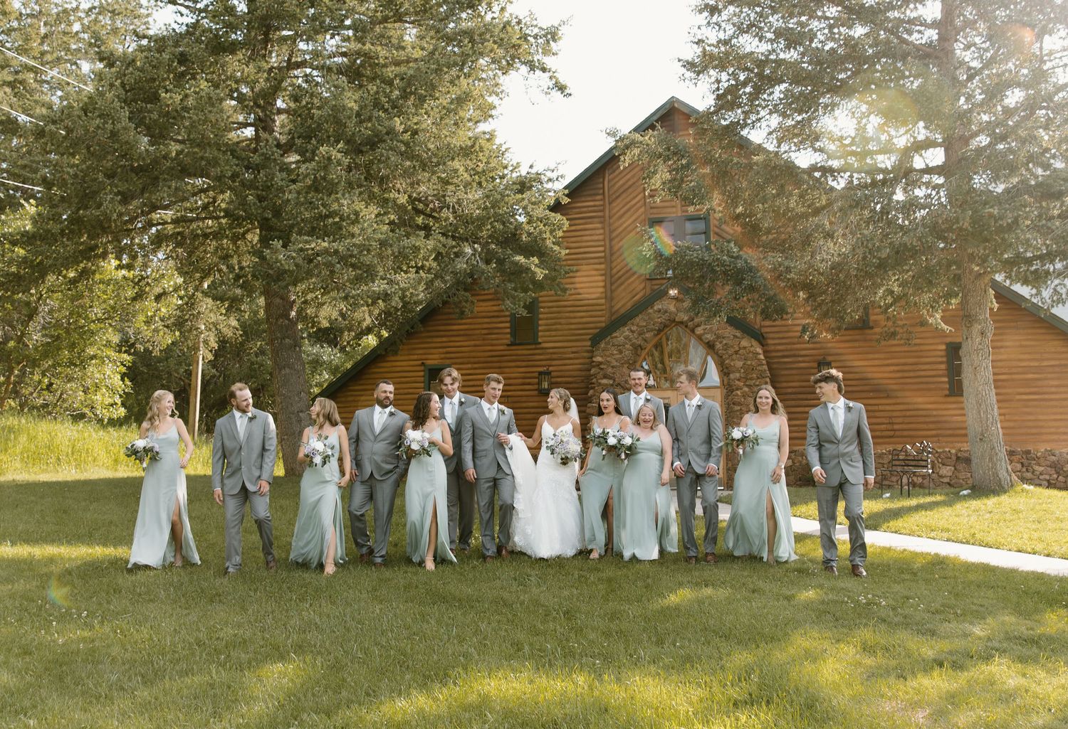 Wedding party in light blue attire poses together in front of a rustic wooden chapel on a sunny afternoon.