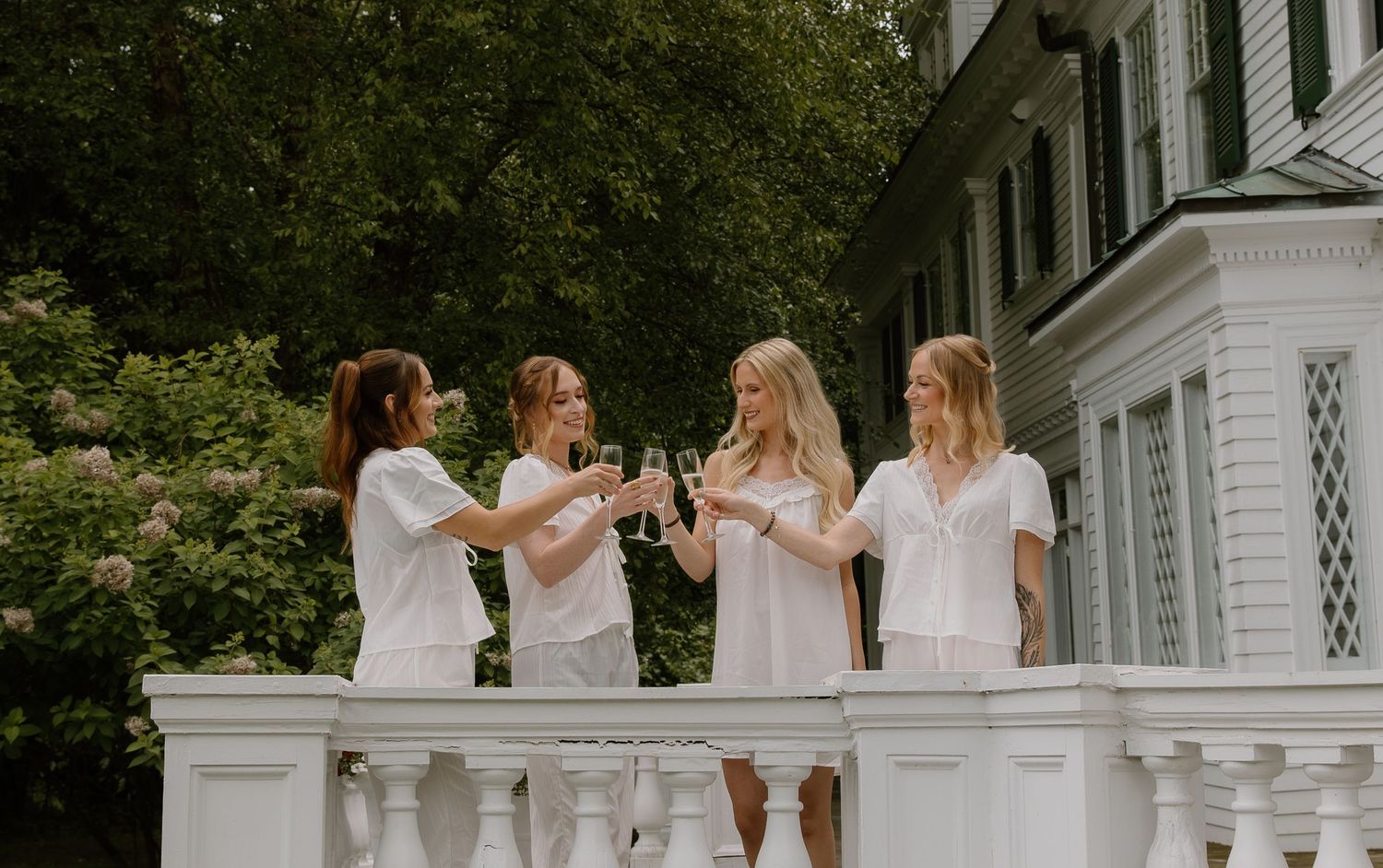 Four women in white dresses toasting with champagne glasses on a elegant white balcony surrounded by lush greenery.