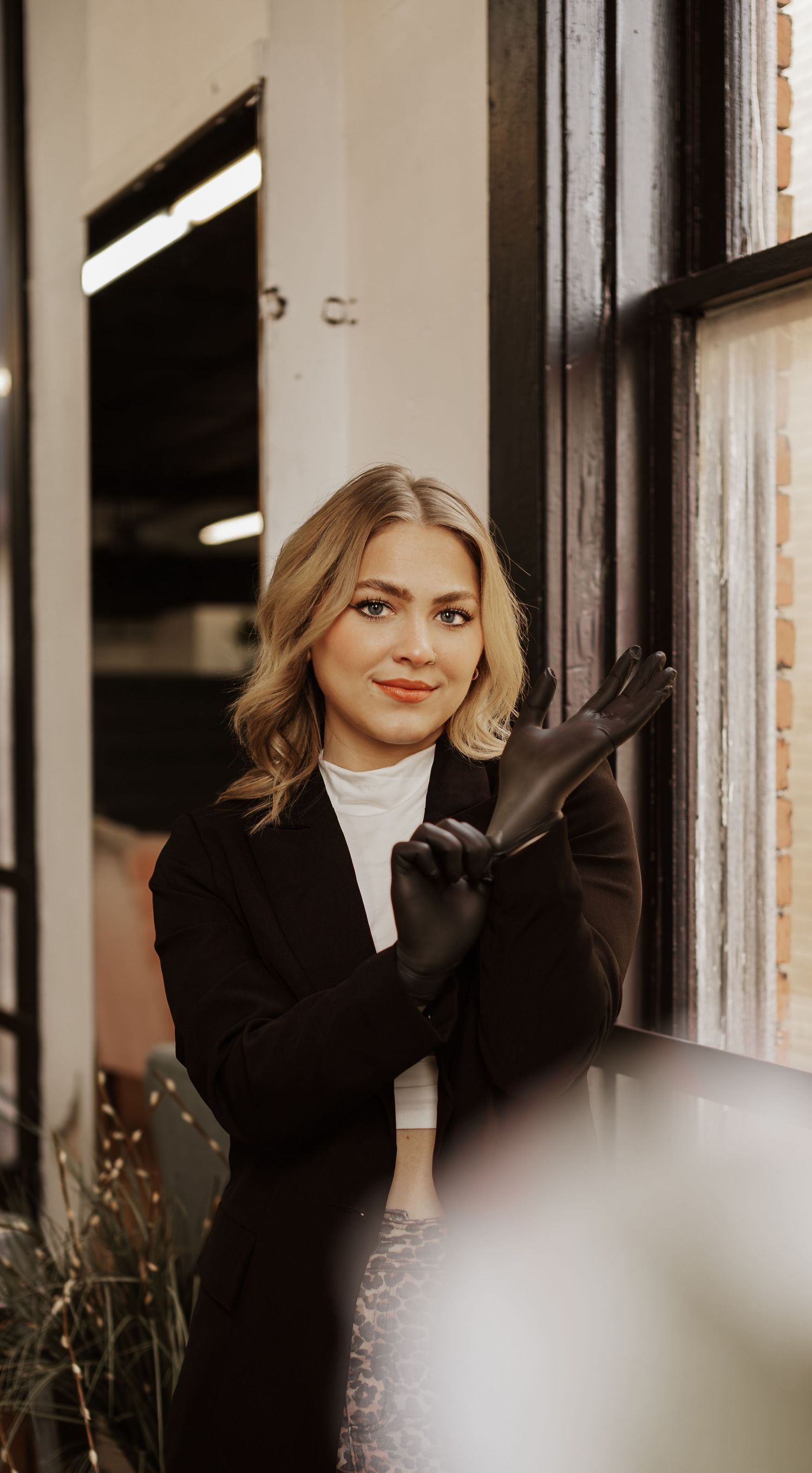 A person wearing a black blazer and leather gloves poses by a window in warm lighting.