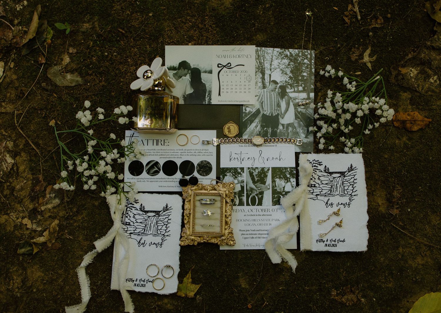Elegant wedding stationery flat lay with invitations, photos, rings, and white flowers on a dark background.