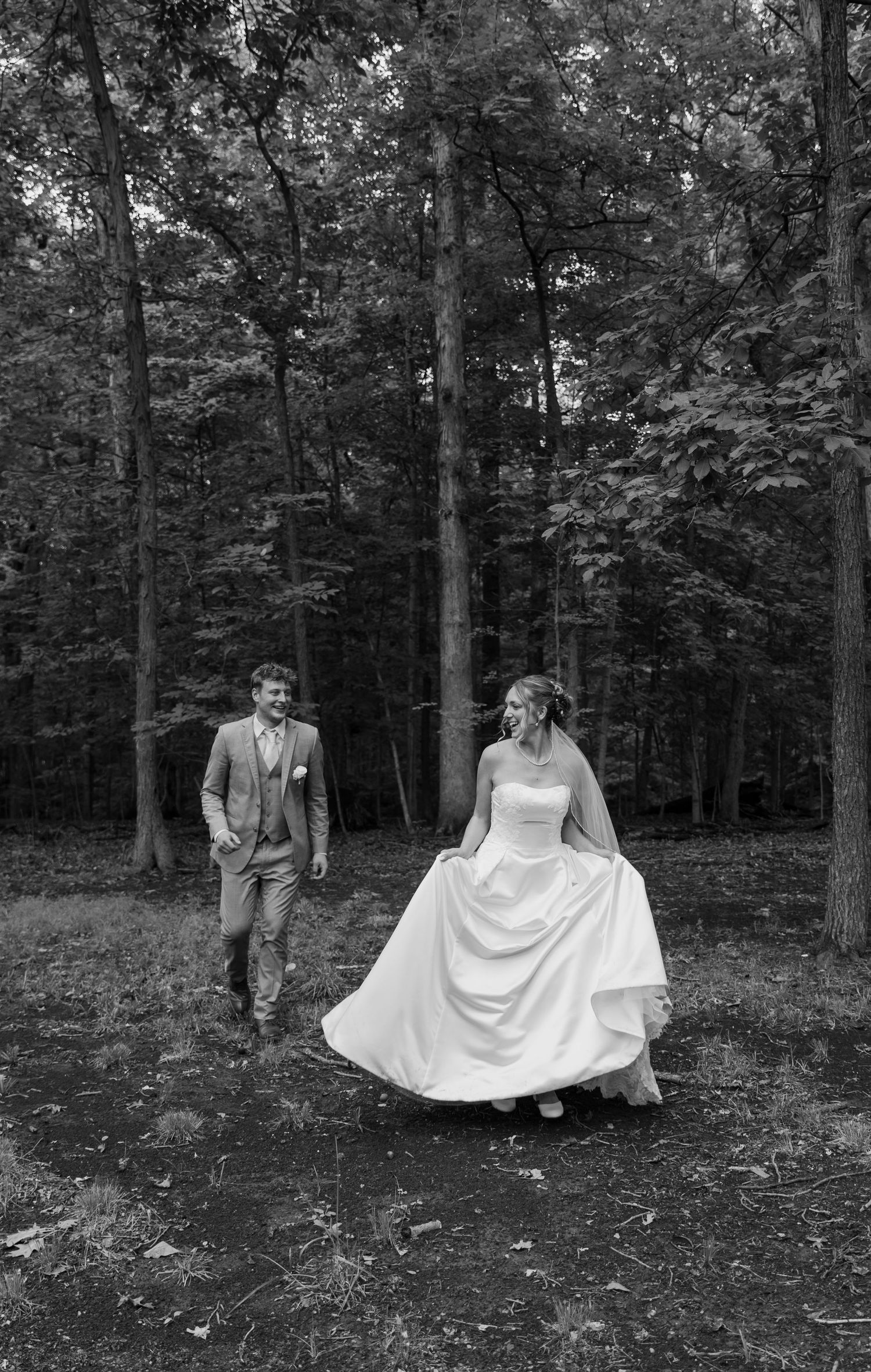 Bride and groom walking through forest in black and white wedding photo, bride holding dress.