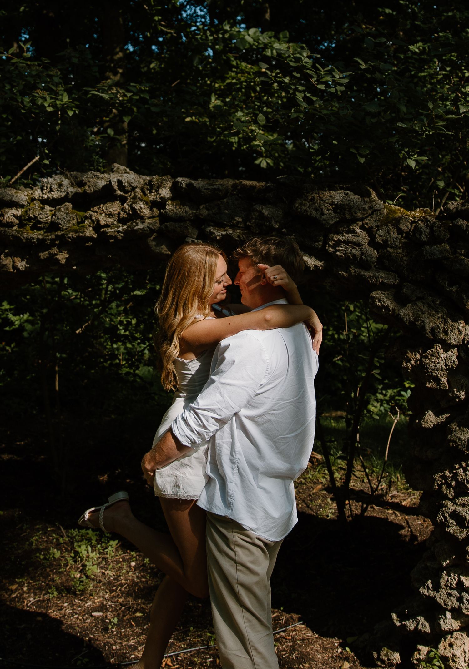 A couple shares a romantic embrace in a sunlit forest, with the man lifting the woman among lush green trees.