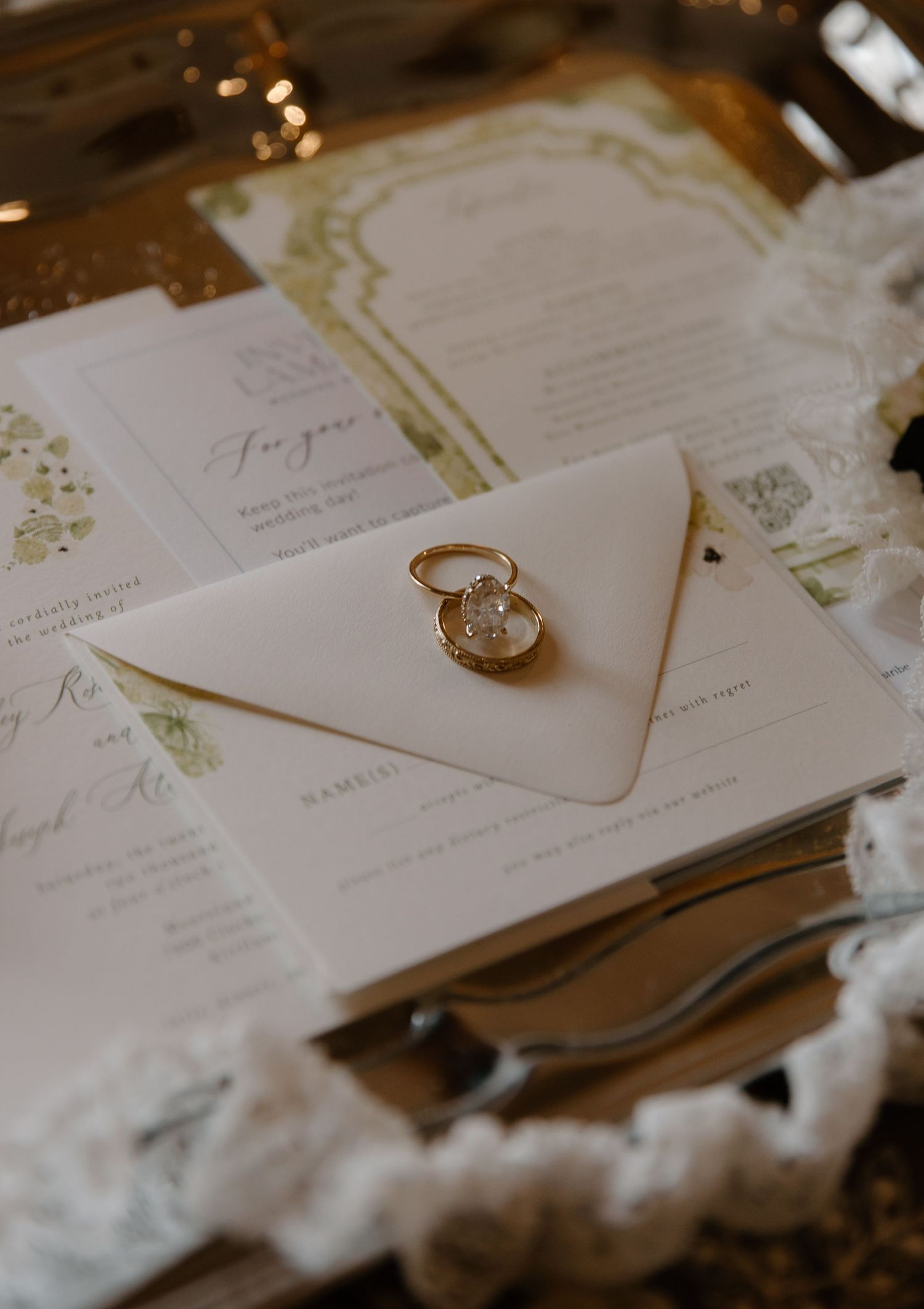 Elegant wedding invitation suite with gold ring on cream envelope, featuring green botanical accents on a gold tray.