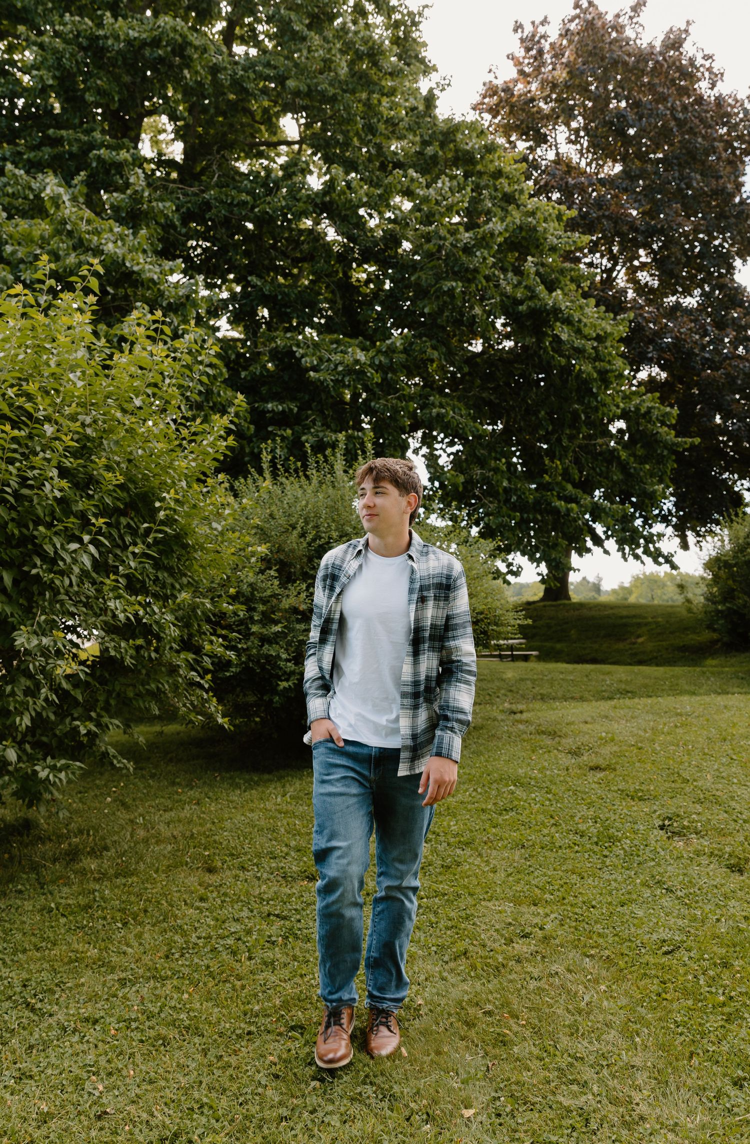 Young man in plaid shirt and jeans standing on green grass with lush trees in background.