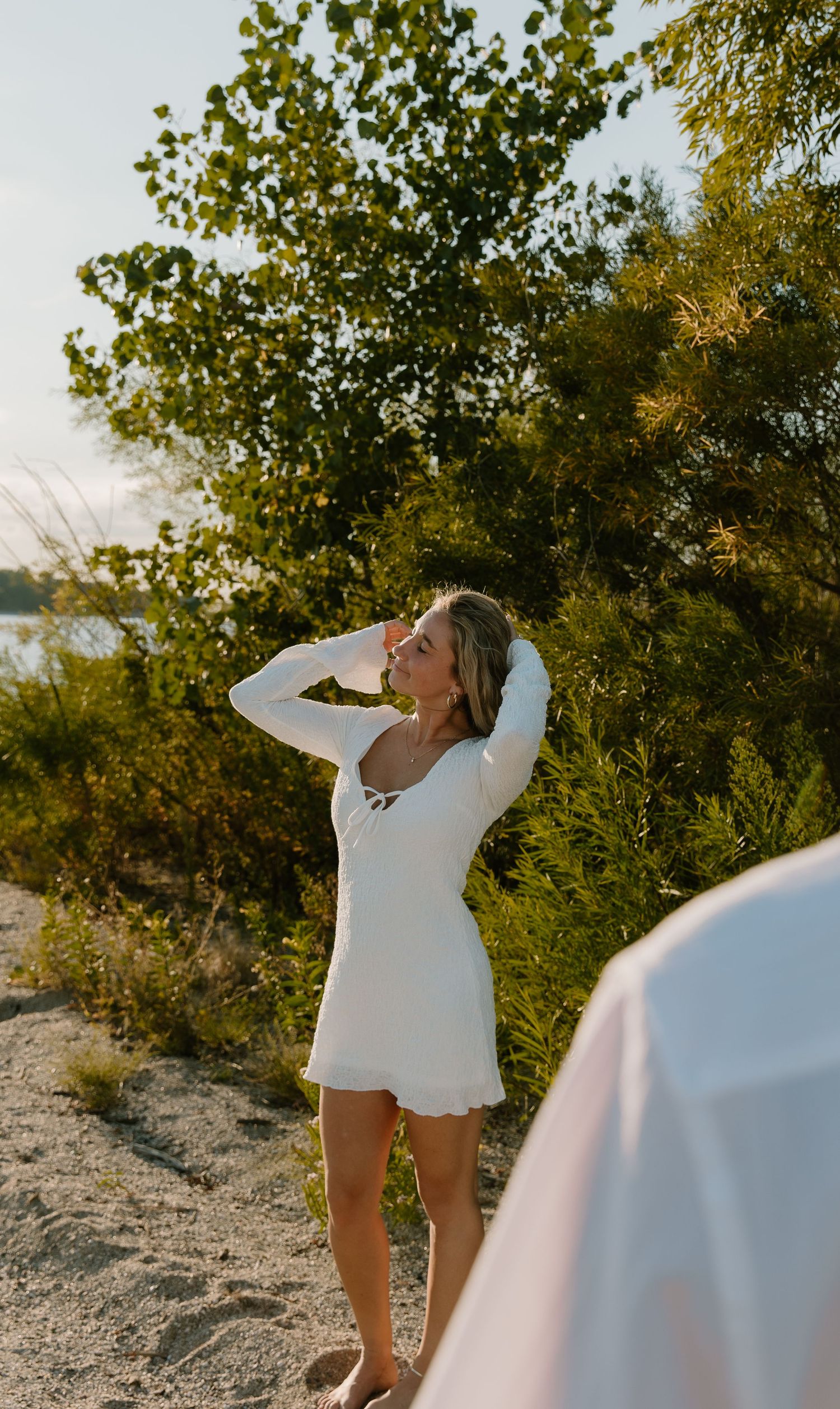 A woman in a white mini dress poses on a sandy beach path surrounded by lush green trees in golden hour light.