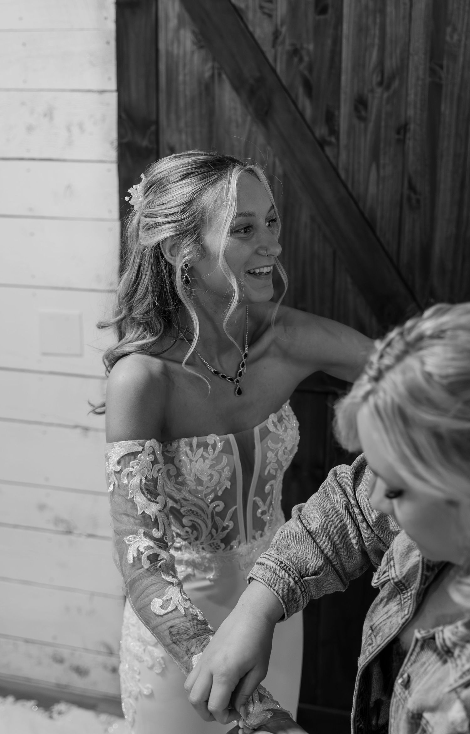Smiling bride in off-shoulder lace wedding gown laughing joyfully near rustic barn doors in black and white photo.