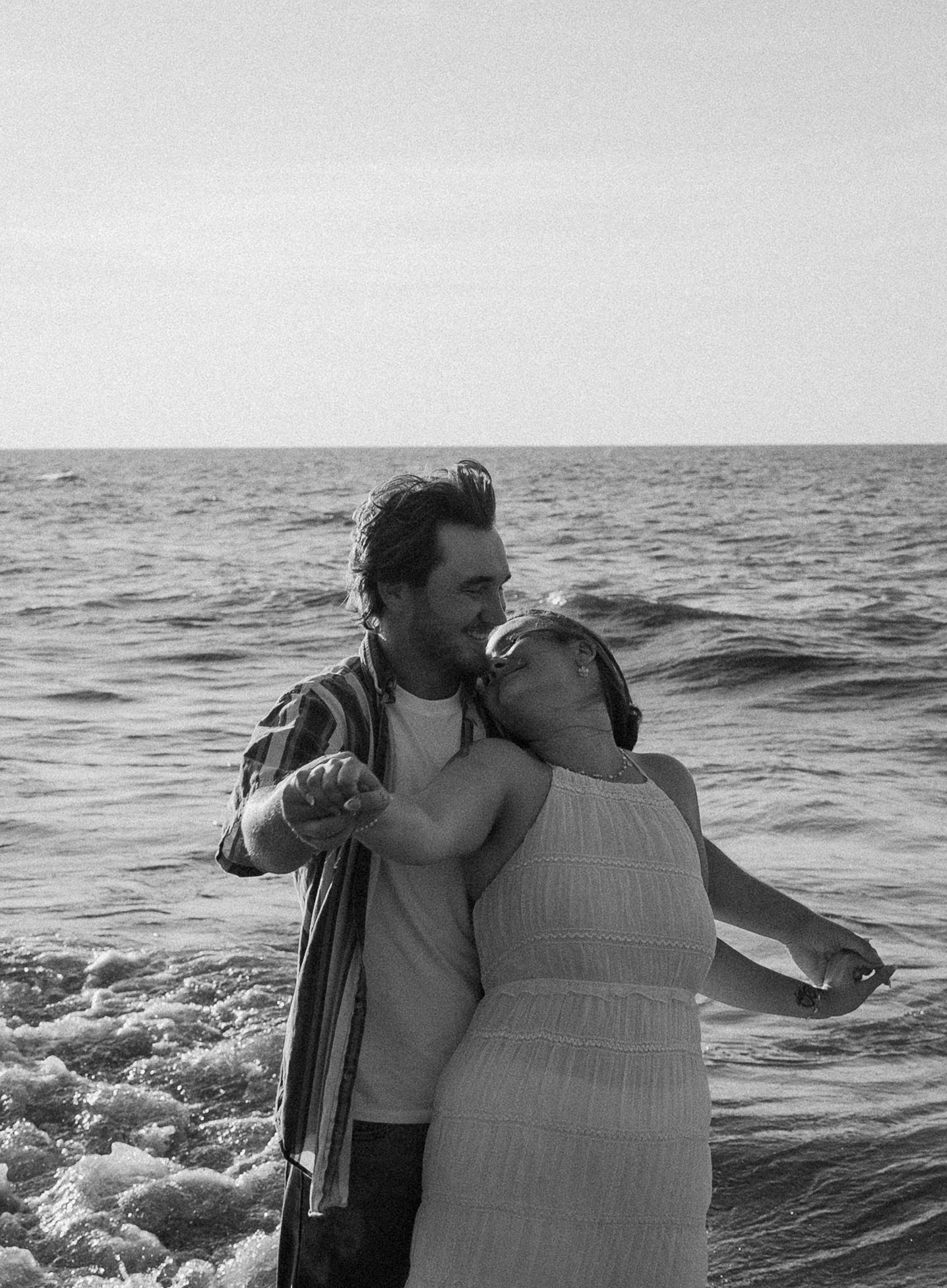 Black and white photo of a romantic couple embracing and dancing together at the ocean shoreline.