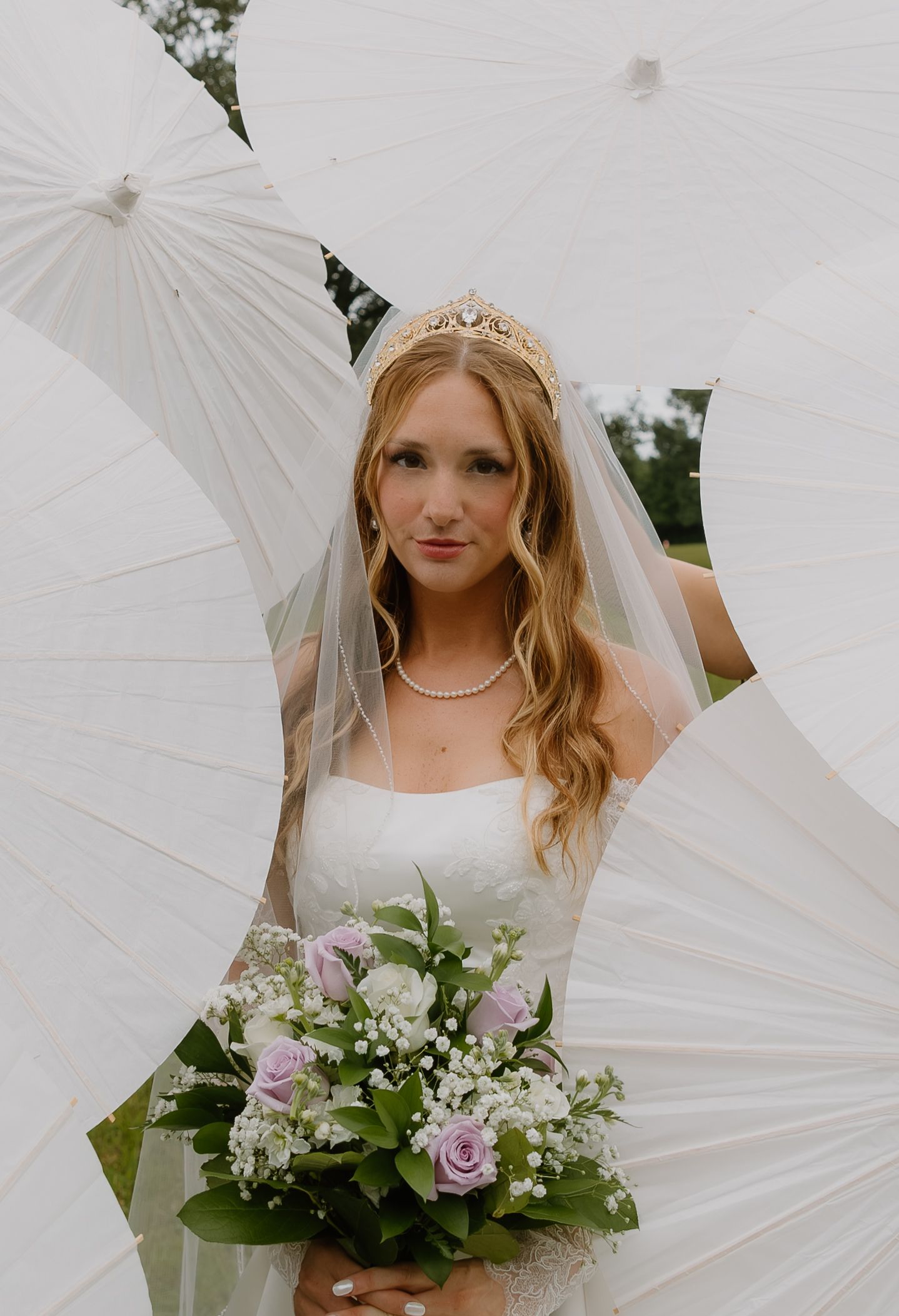 Bride in white gown and tiara holds purple and white bouquet, surrounded by white parasols outdoors.
