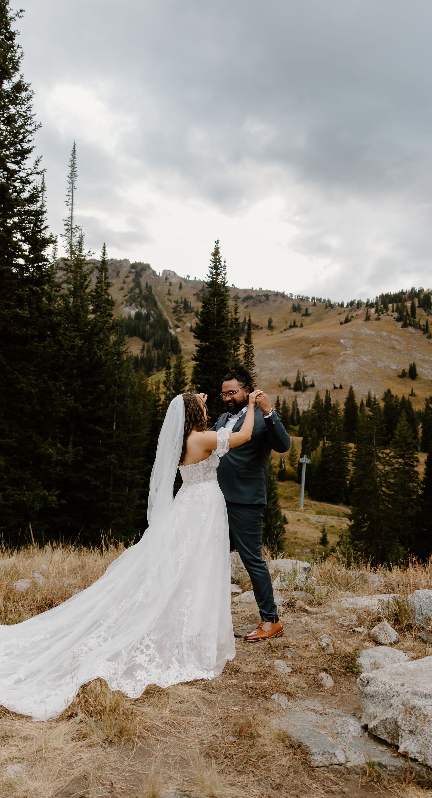 A couple in wedding attire sharing a romantic moment on a mountain trail with evergreen trees and peaks in the background.