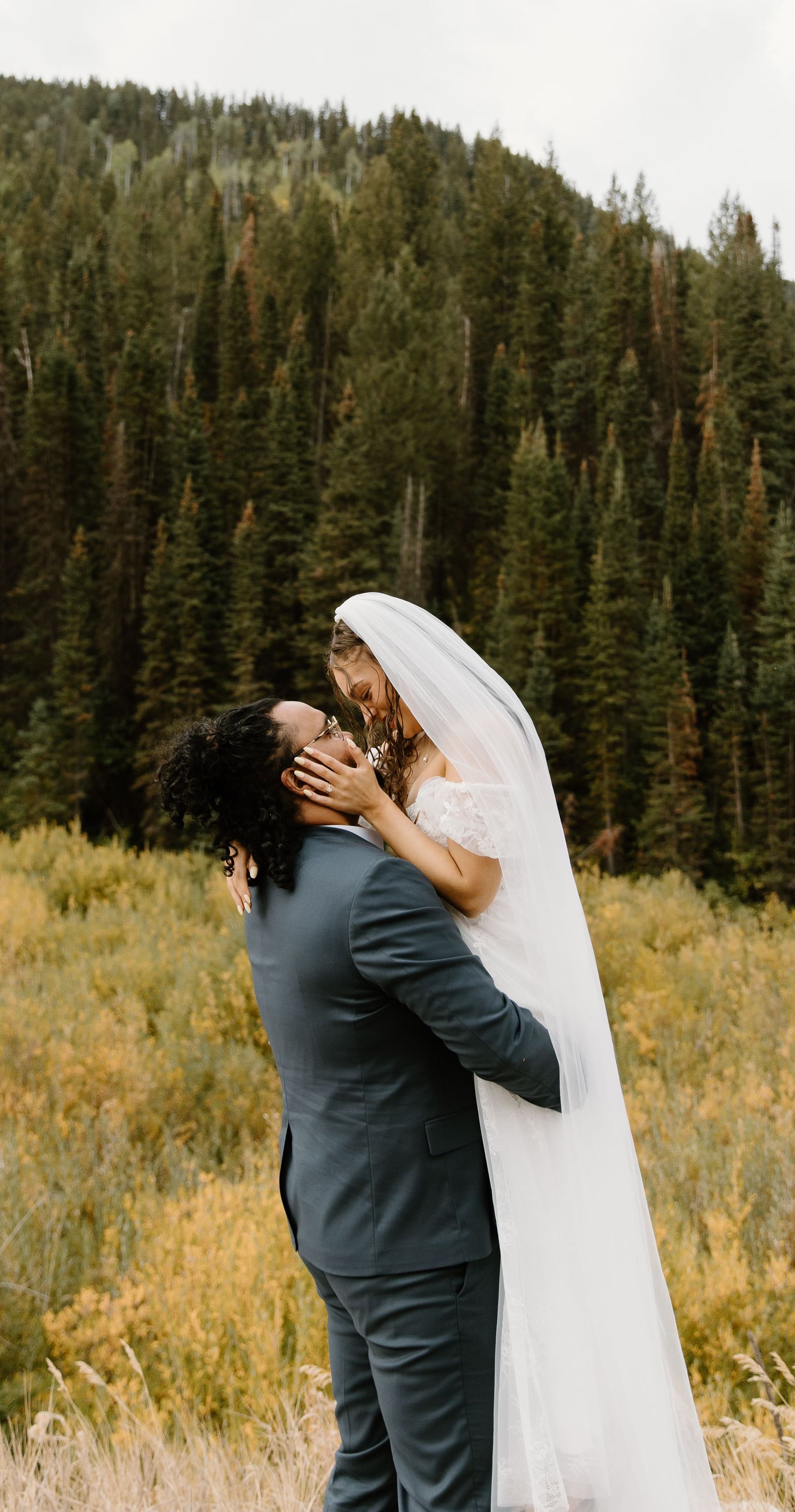A couple embraces in a romantic kiss against a backdrop of evergreen forest and golden meadow.