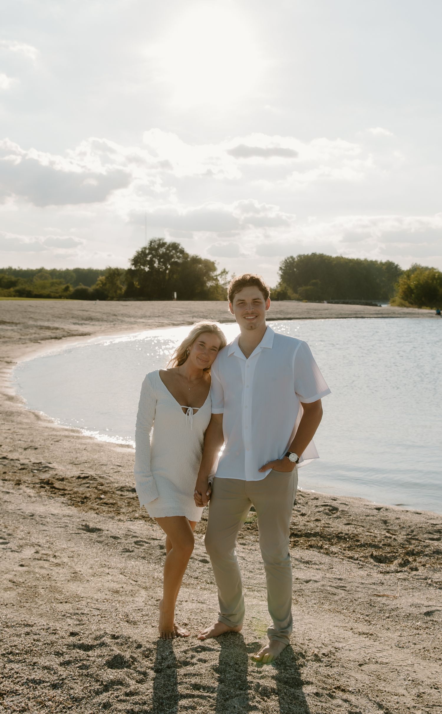 Couple holding hands on sandy beach at golden hour, woman in white dress, man in light blue shirt and khaki pants.