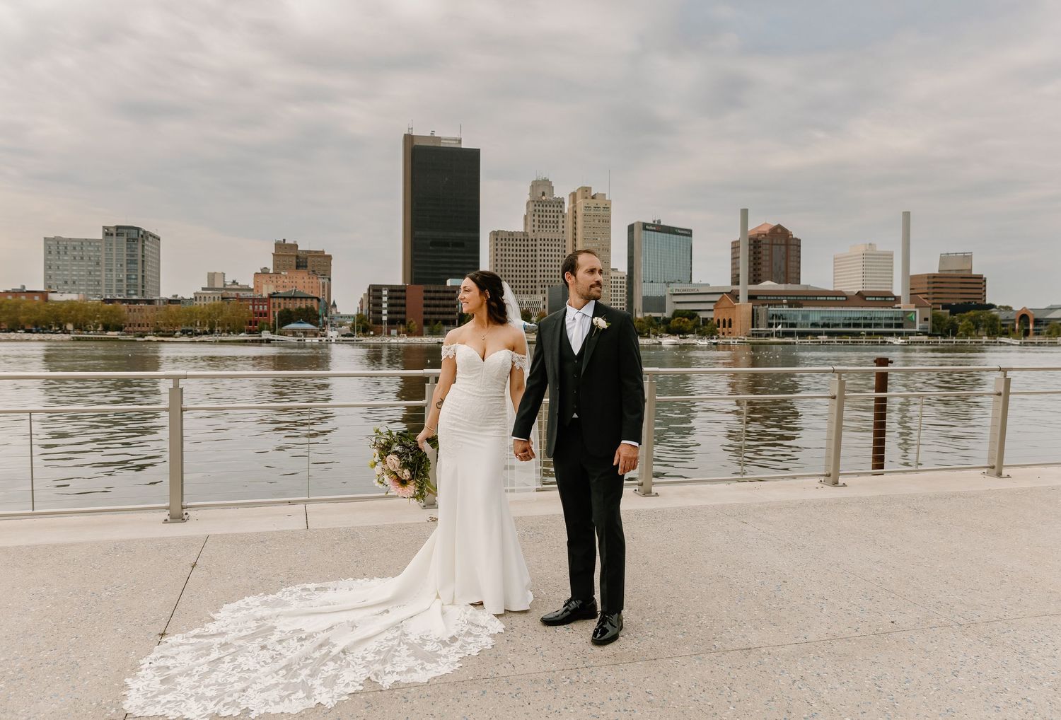 A couple in wedding attire walks along a waterfront promenade with a city skyline in the background.