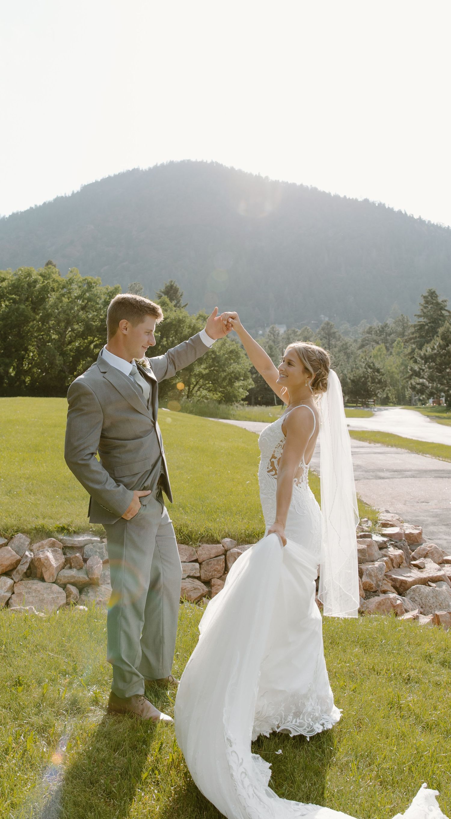 A bride and groom dance playfully together on a grassy hillside with mountains in the background.