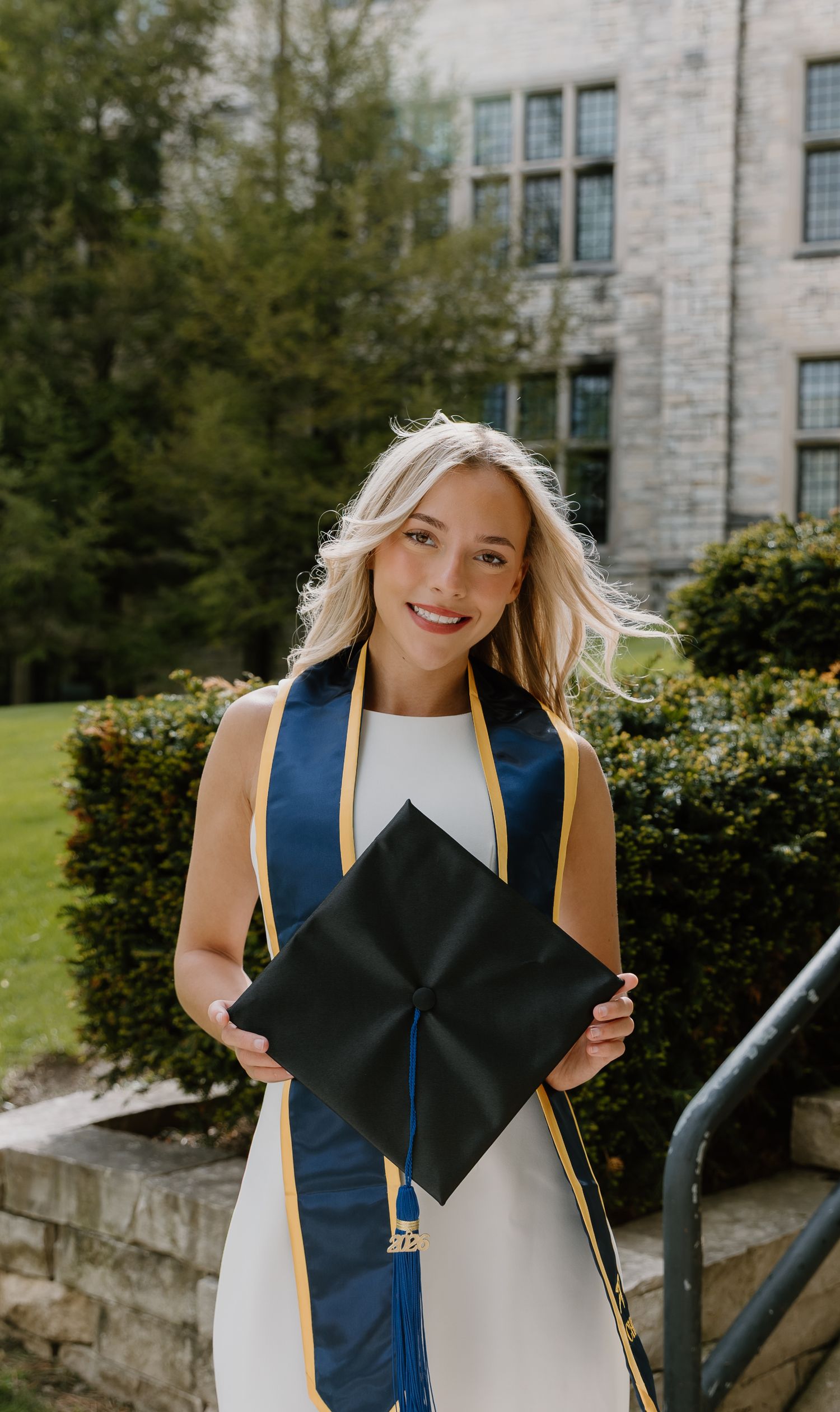 Smiling blonde graduate holding black mortarboard cap, wearing navy and gold sash over white dress outdoors.
