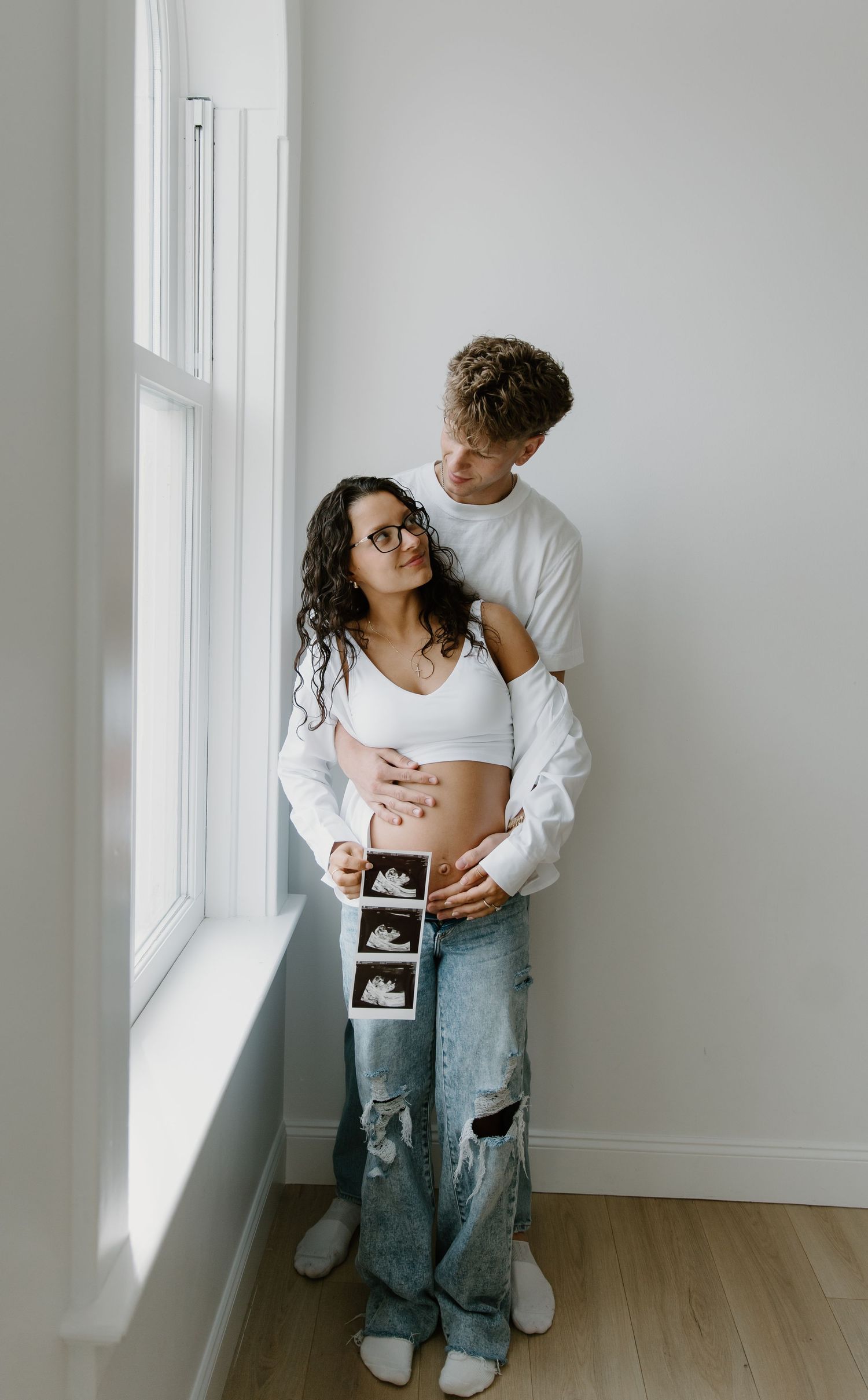 Pregnant couple holding ultrasound photos by window, woman in white crop top and ripped jeans, man embracing her from behind.