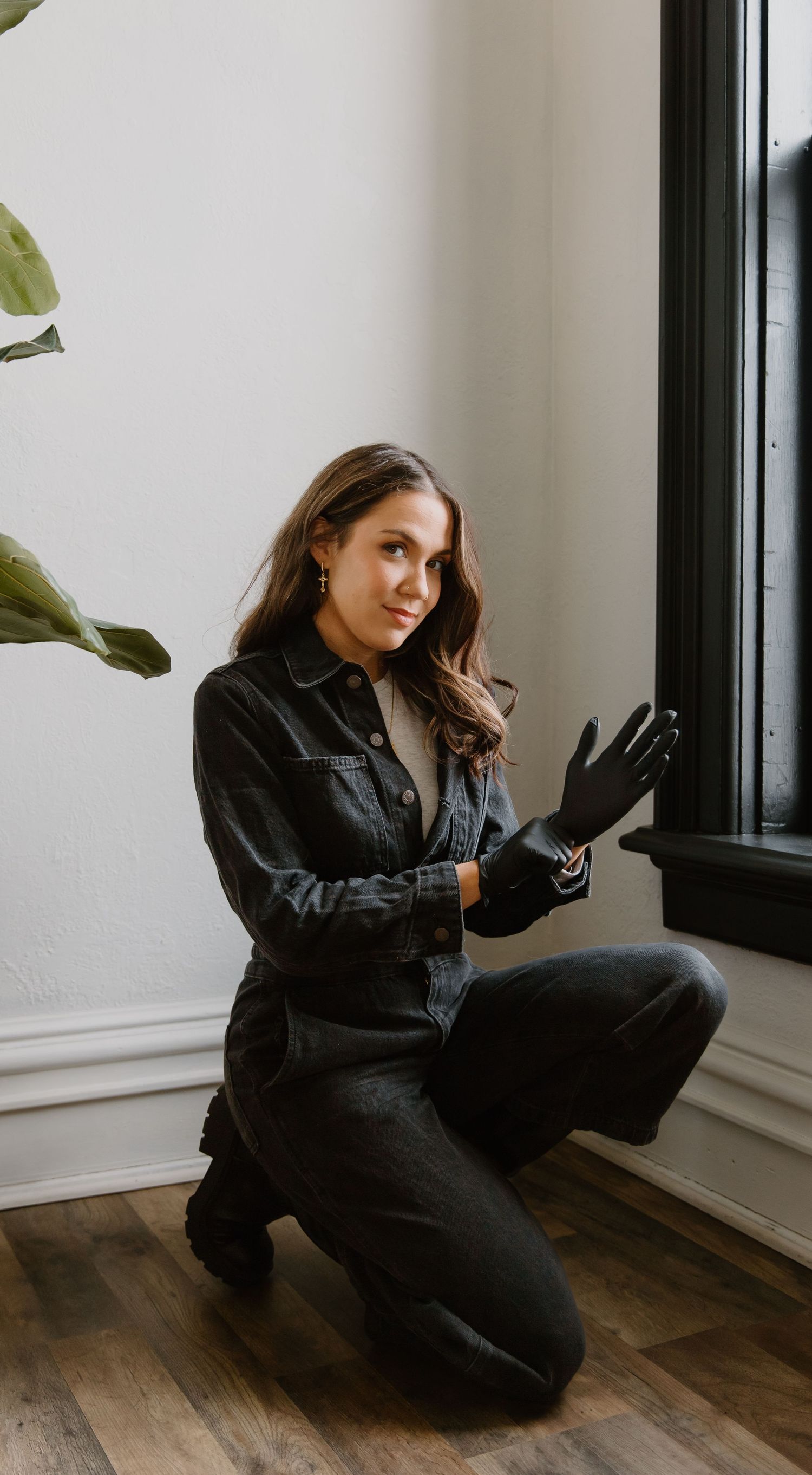 Woman in dark denim outfit and black gloves kneeling by a window on hardwood floor near a green plant.