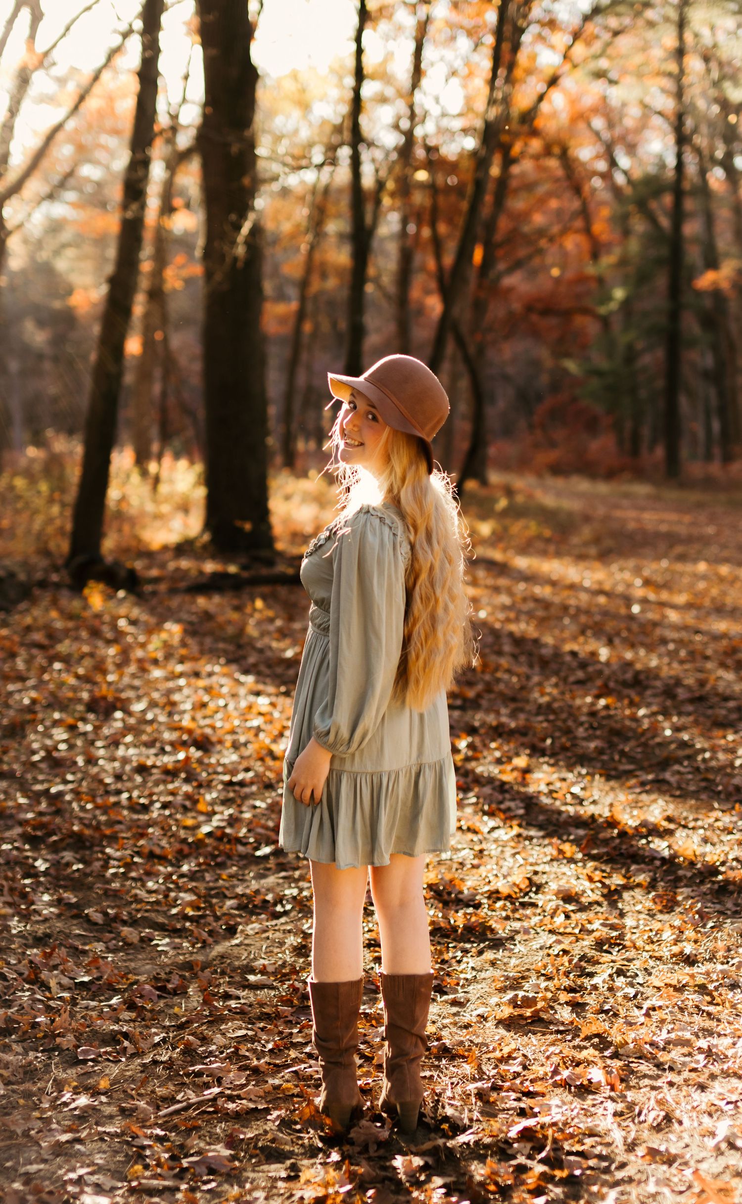 Figure in a flowing dress and boots stands among fallen autumn leaves in a sunlit forest.