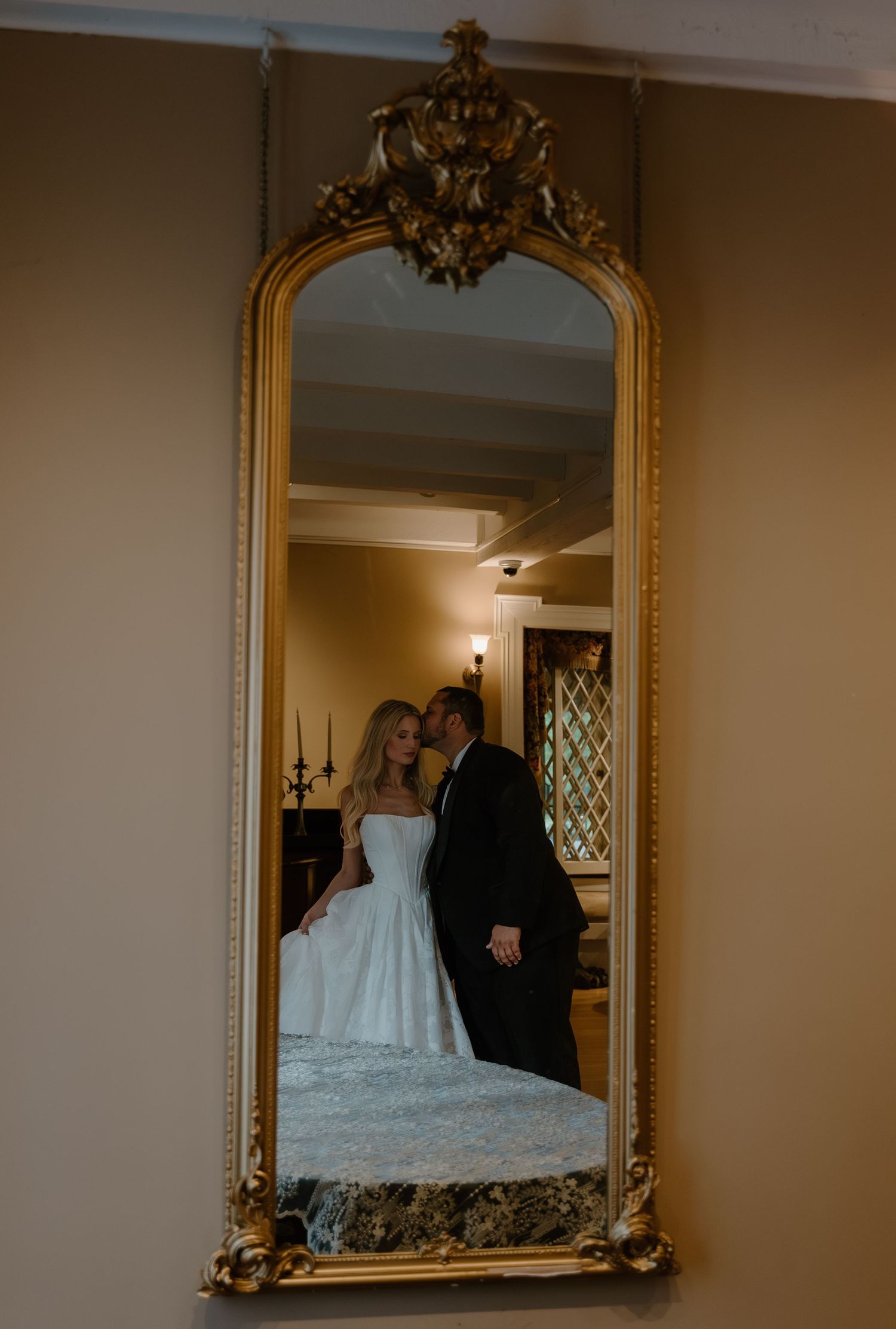 Bride and groom share a romantic moment reflected in an ornate gold-framed mirror in elegant venue.