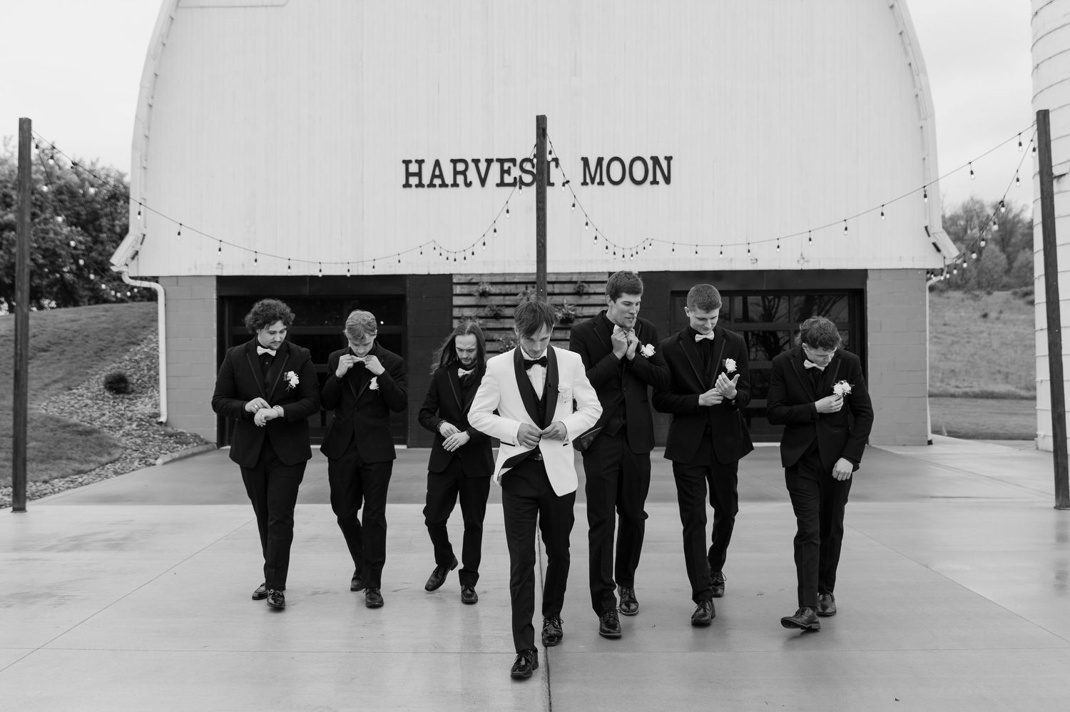 Groom in white tuxedo jacket walks with groomsmen in black suits outside Harvest Moon wedding venue.