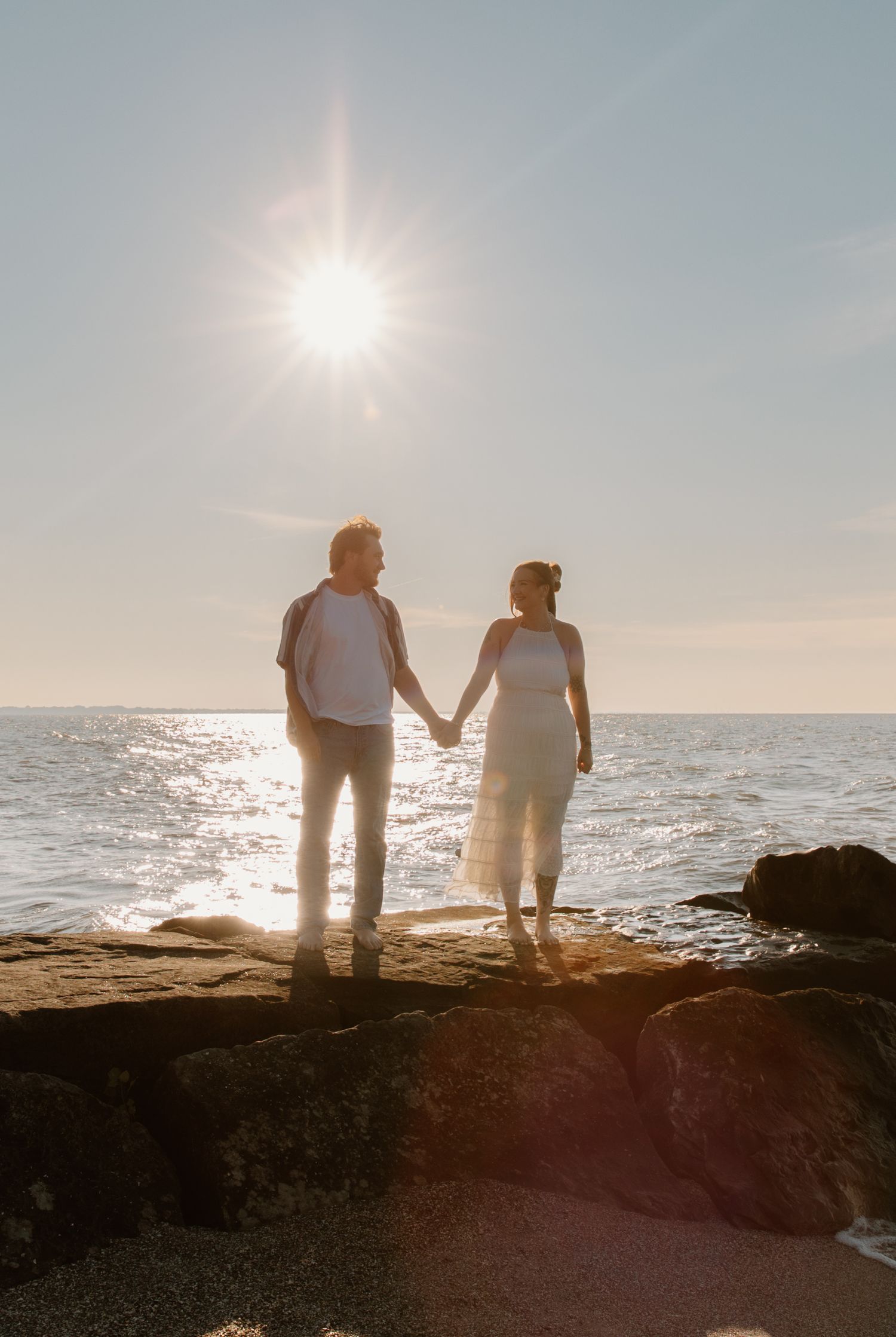 Couple holding hands on rocky shore at sunset with sun flare over sparkling ocean in background.