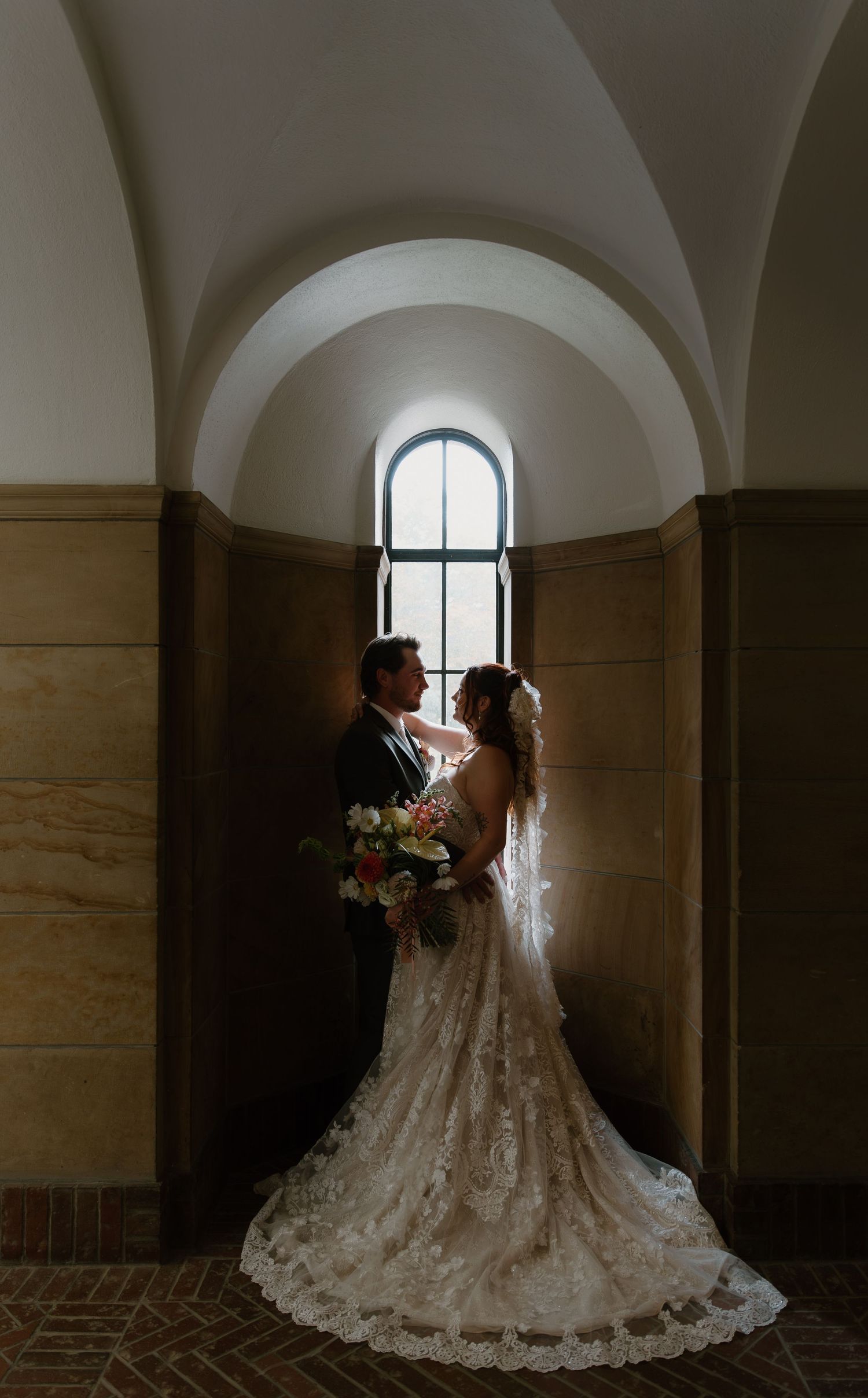 Bride and groom share a romantic moment in an arched hallway, bride holding a colorful bouquet in a lace gown.