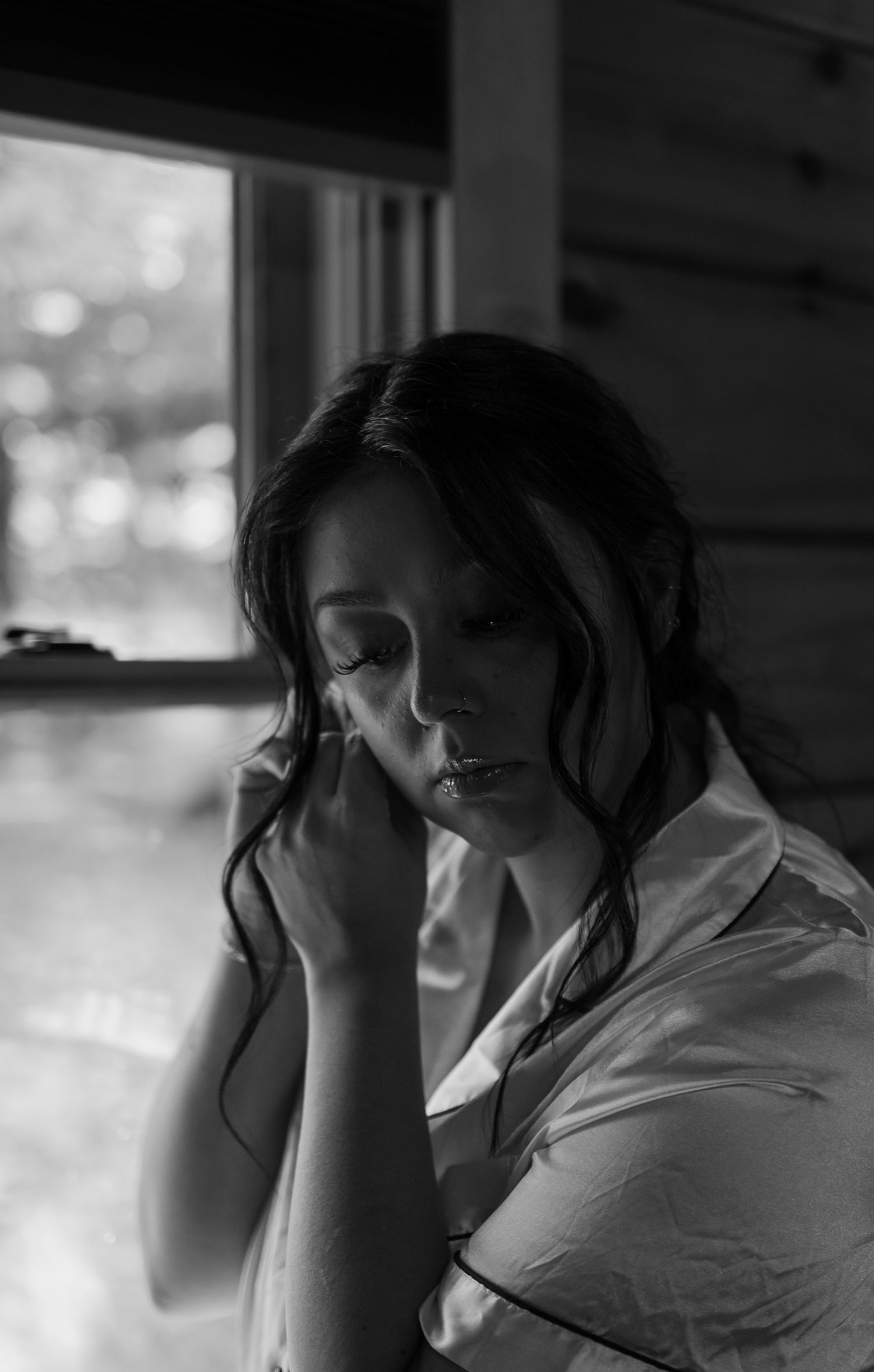 Black and white portrait of a pensive woman with dark hair, eyes closed, hand raised to her face near a window.