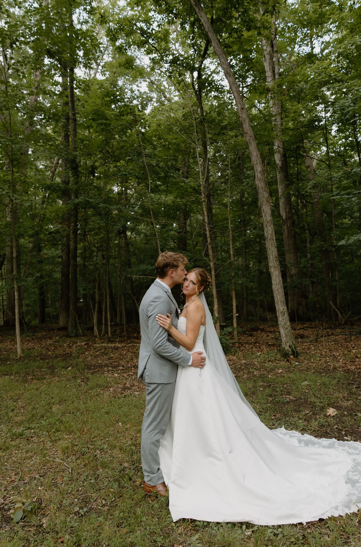A bride and groom share a romantic kiss in a lush forest setting, her white gown and veil trailing gracefully on the ground.