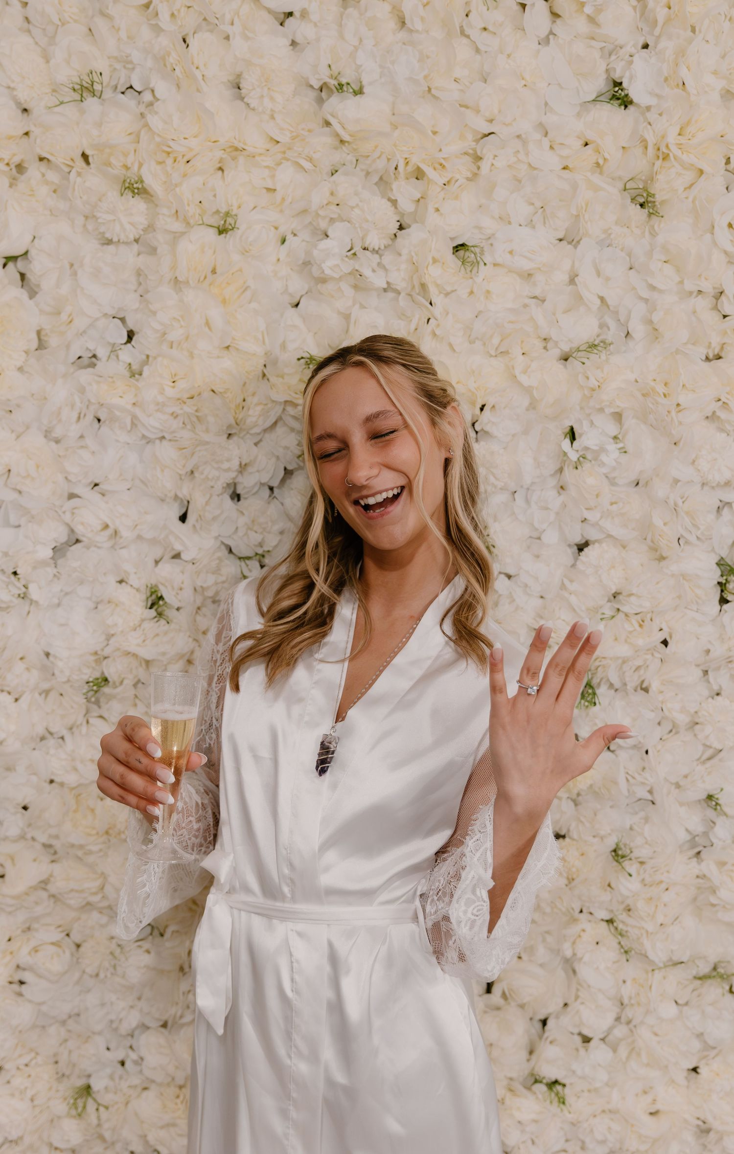 A laughing bride in a white lace robe holds champagne and waves in front of a lush white floral wall backdrop.