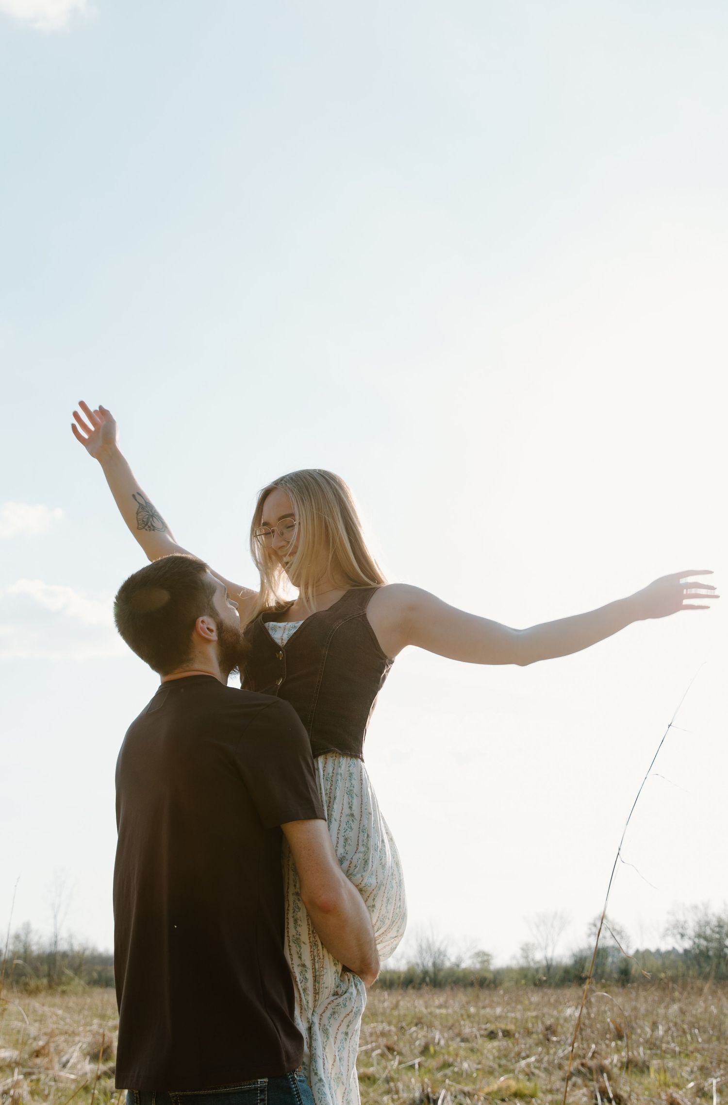 A man lifts a woman with outstretched arms in a sunny field, capturing a romantic outdoor moment.