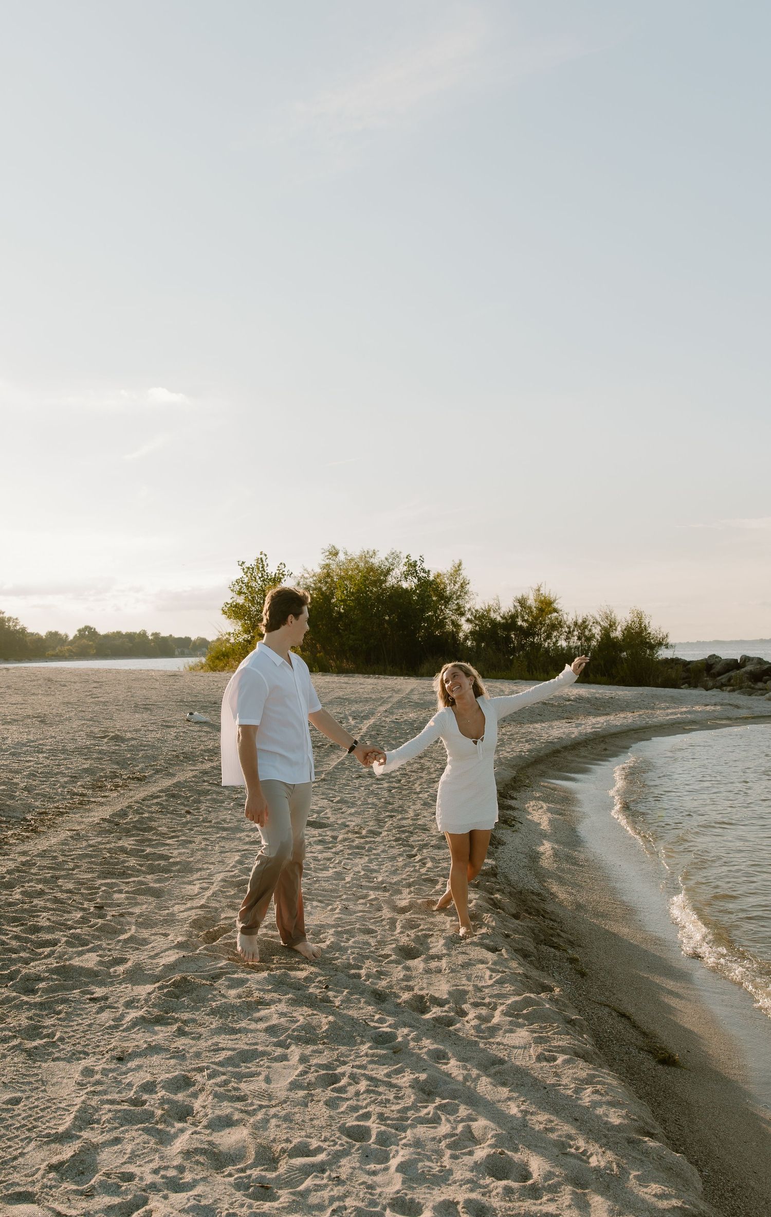 Couple holding hands and dancing on a sandy beach at golden hour, woman in white dress with arm raised.