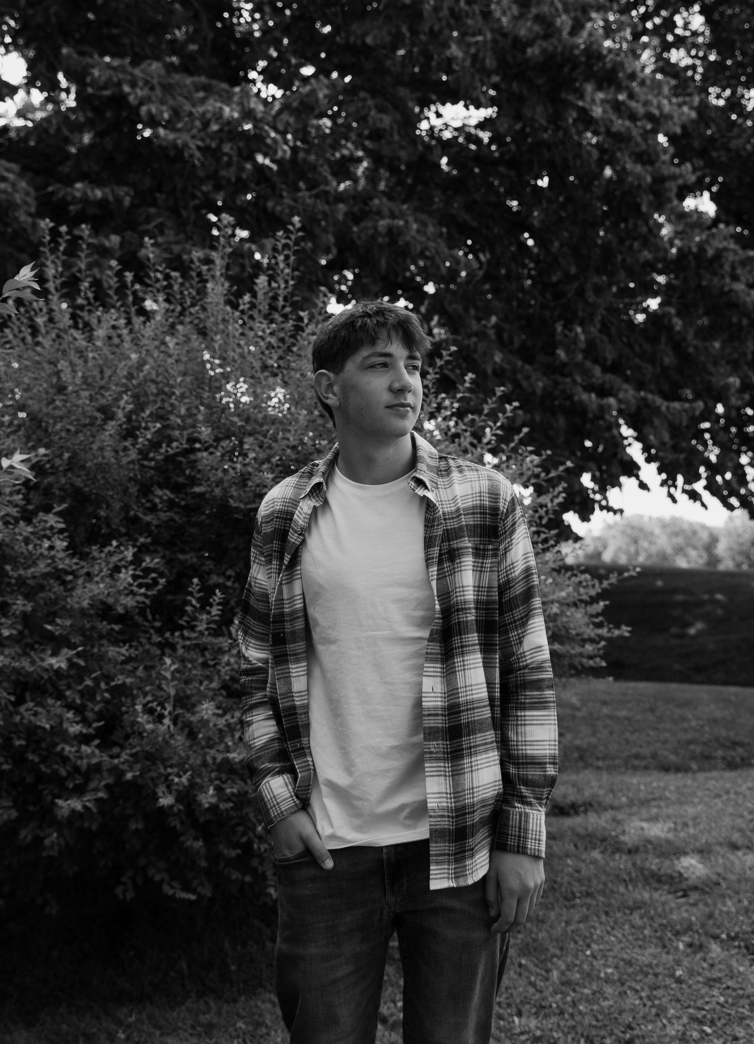 Young man in plaid flannel shirt and jeans standing outdoors near green trees in black and white photo.