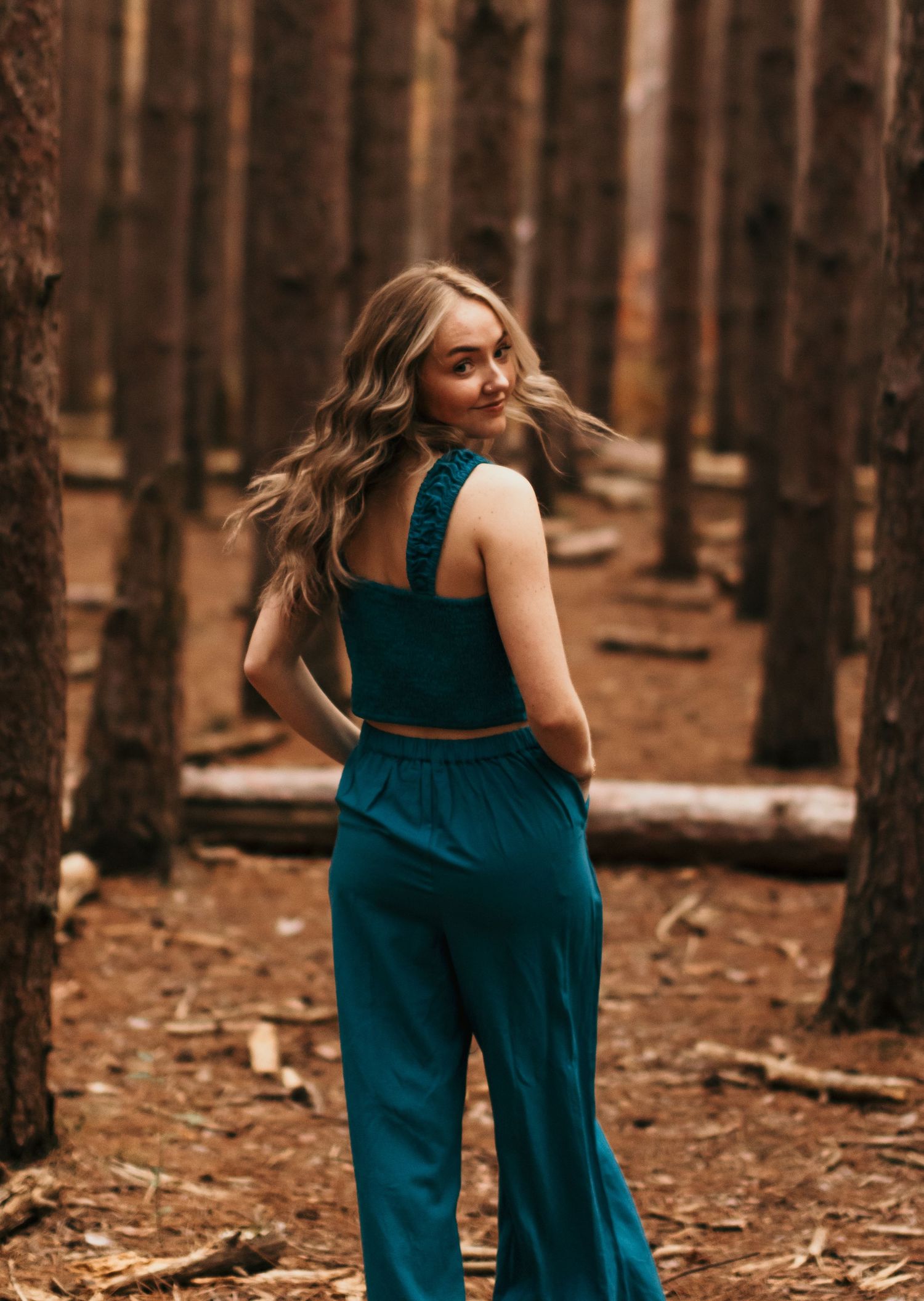 A woman in a teal jumpsuit poses in a forest setting with warm autumn tones.