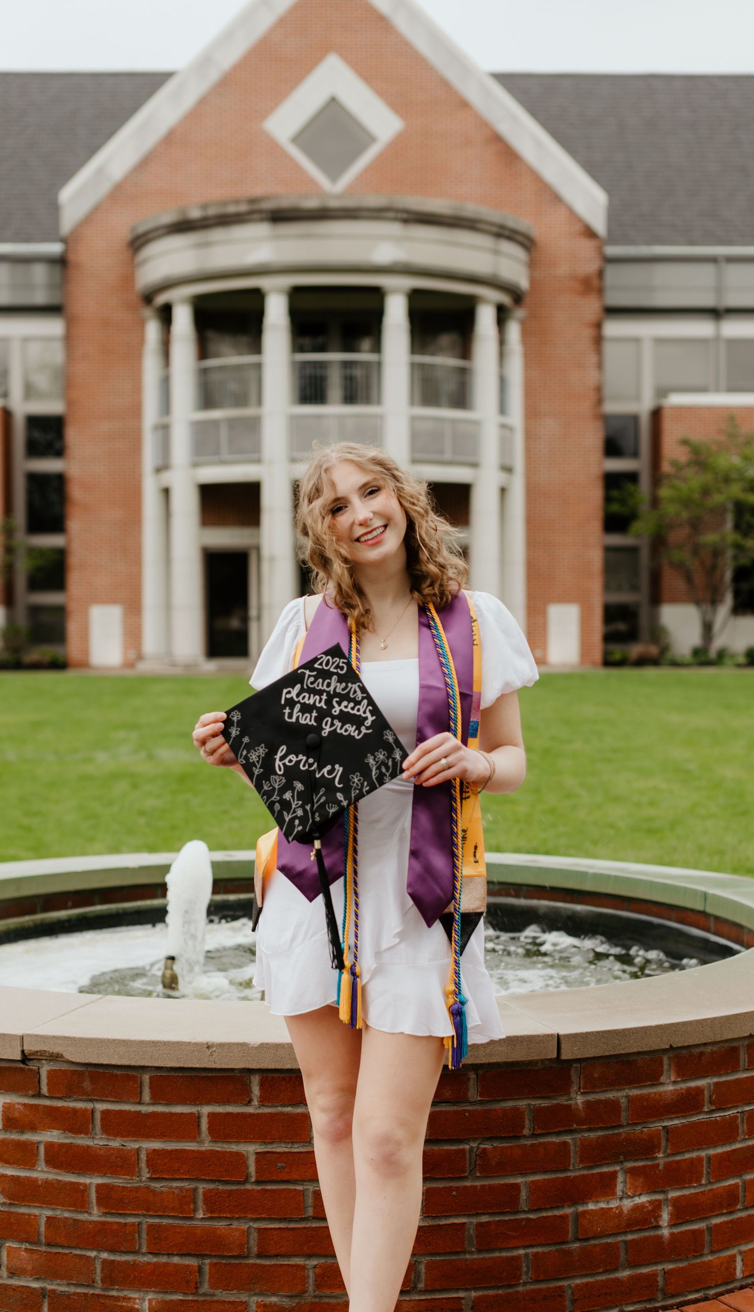 Graduate wearing a purple stole and white dress poses with decorated cap in front of a brick building with fountain.